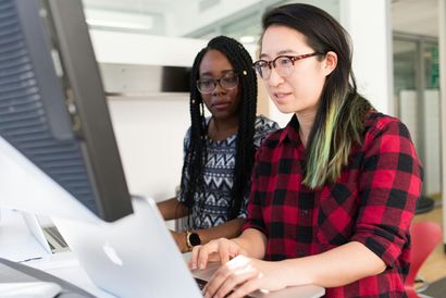 Two women looking at a laptop and a computer monitor in a bright office. One types.