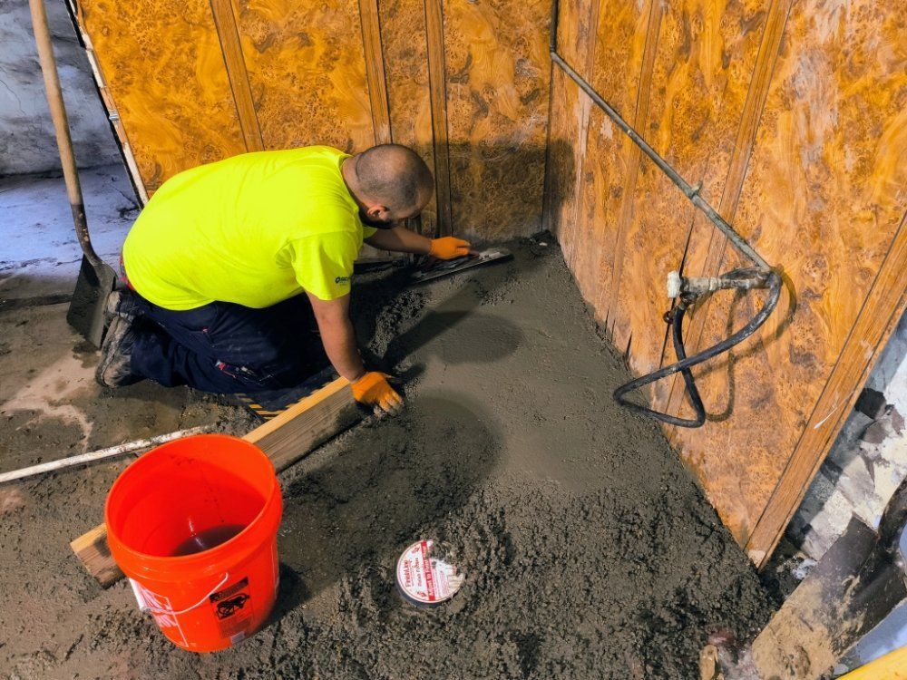 Worker troweling wet concrete beside a plywood wall, with an orange bucket in the foreground.