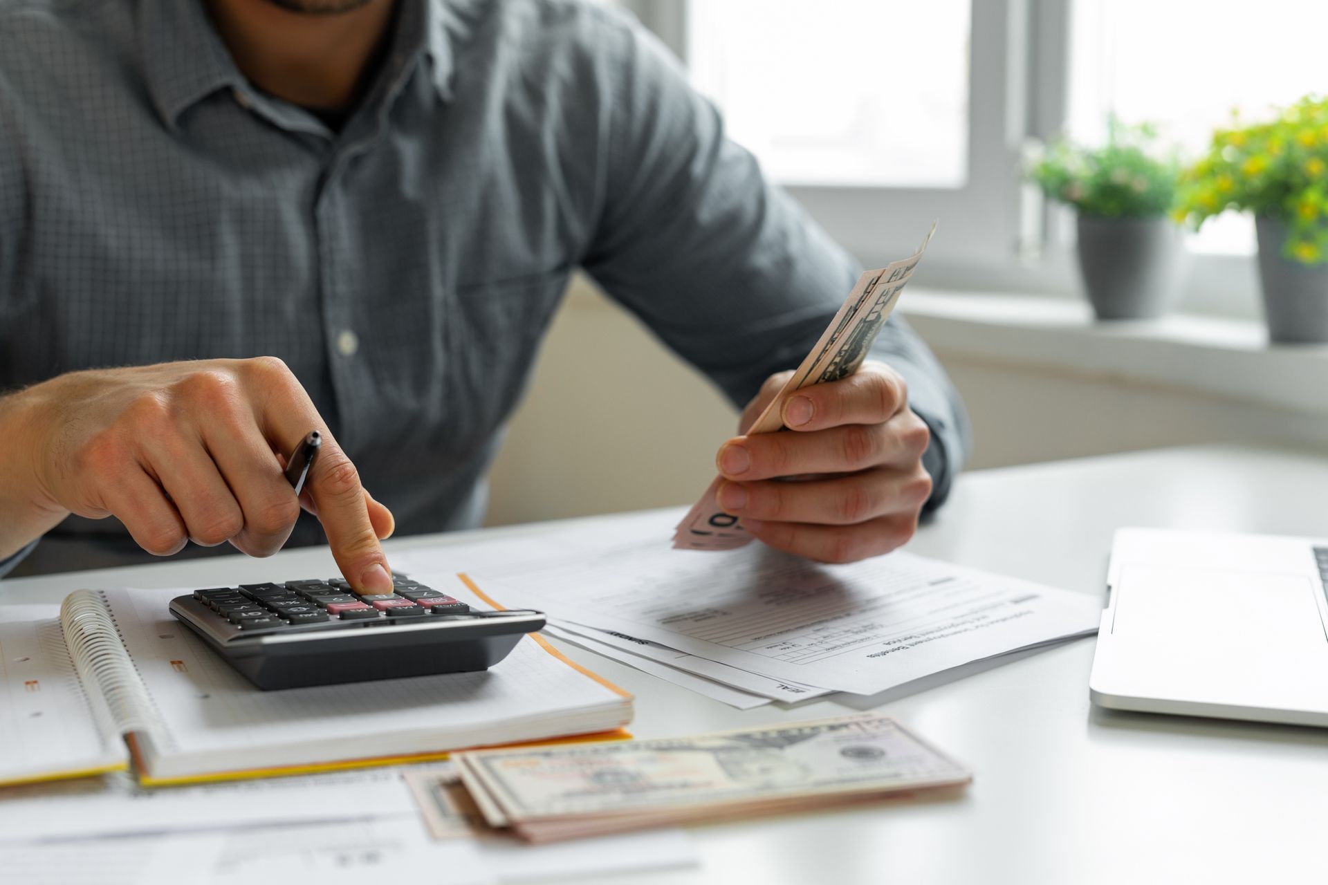 A man is sitting at a table using a calculator and holding a stack of money.