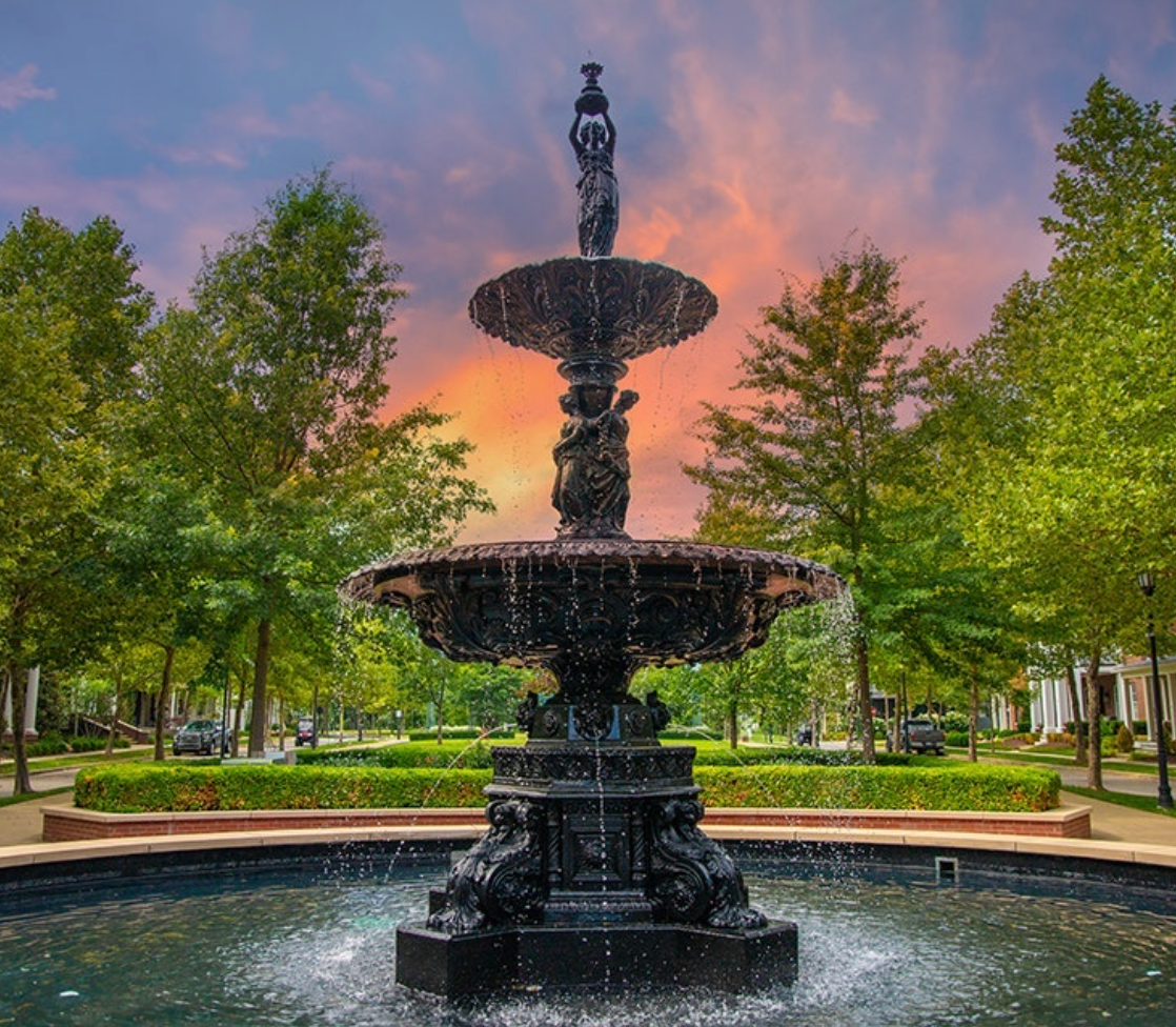 Ornate fountain in a pool with water spraying, surrounded by trees and a colorful sunset.