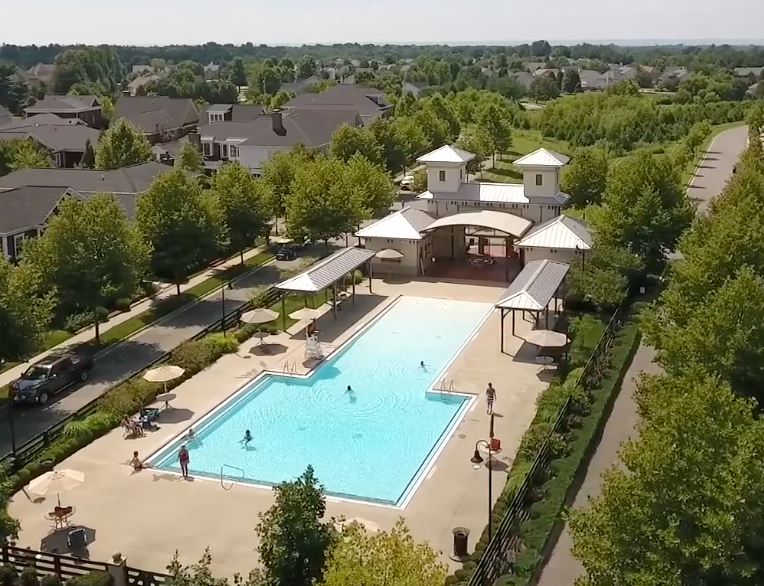 Aerial view of a rectangular swimming pool with people in the water, surrounded by concrete, trees, and buildings. Purple Martin Hotel