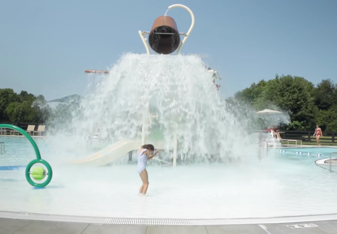 Child in a swimsuit stands in a shallow pool as a large bucket dumps water over them. Purple Martin Hotel