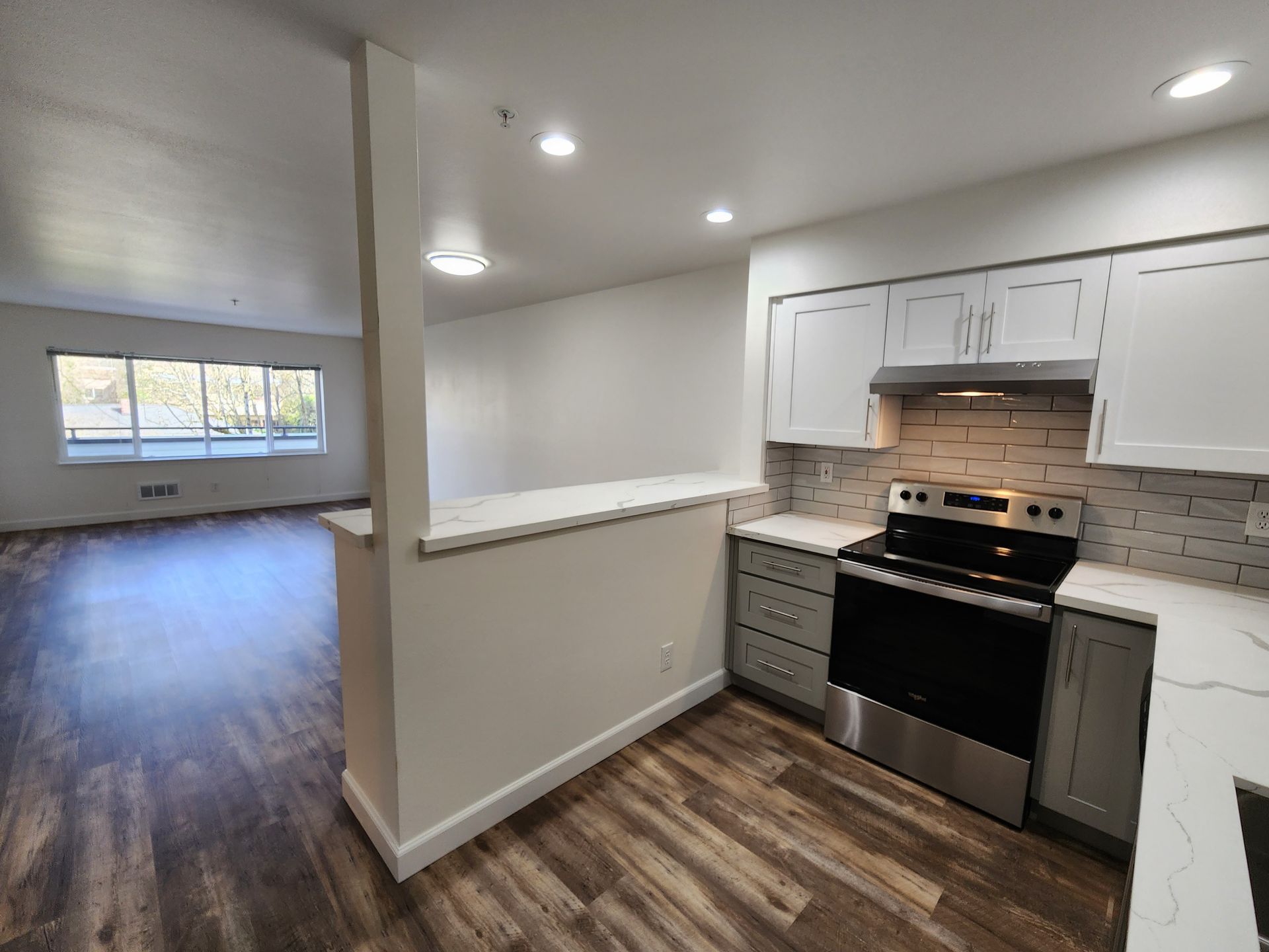 Kitchen with light gray cabinets, stainless steel stove, and open view to living room with large window.