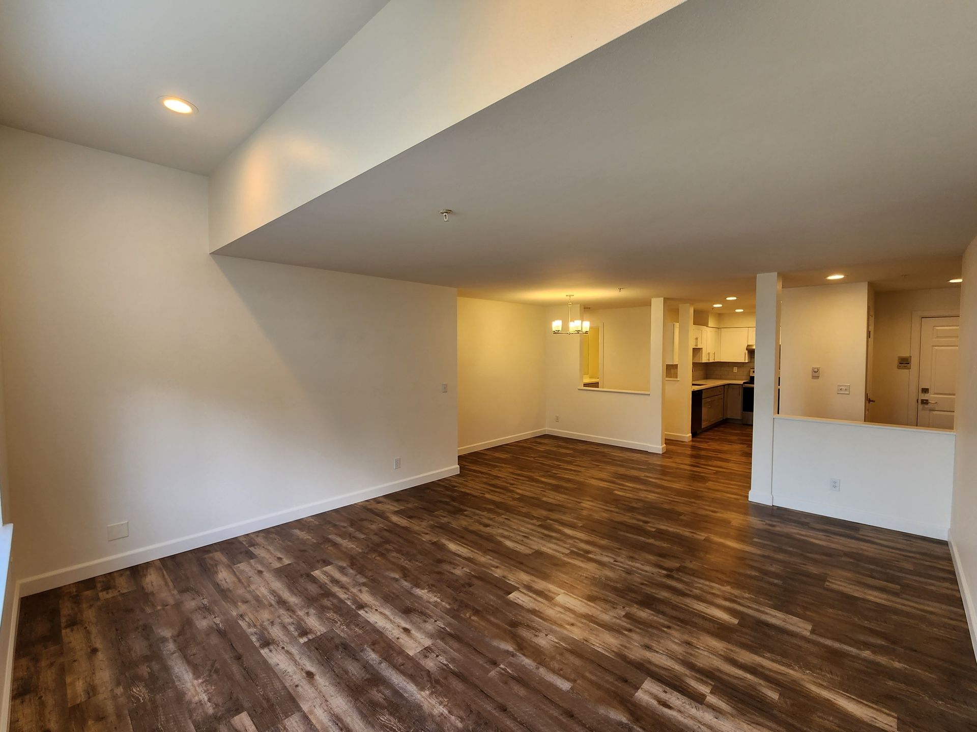 Empty living room with wood-look flooring, white walls, and view into a kitchen.