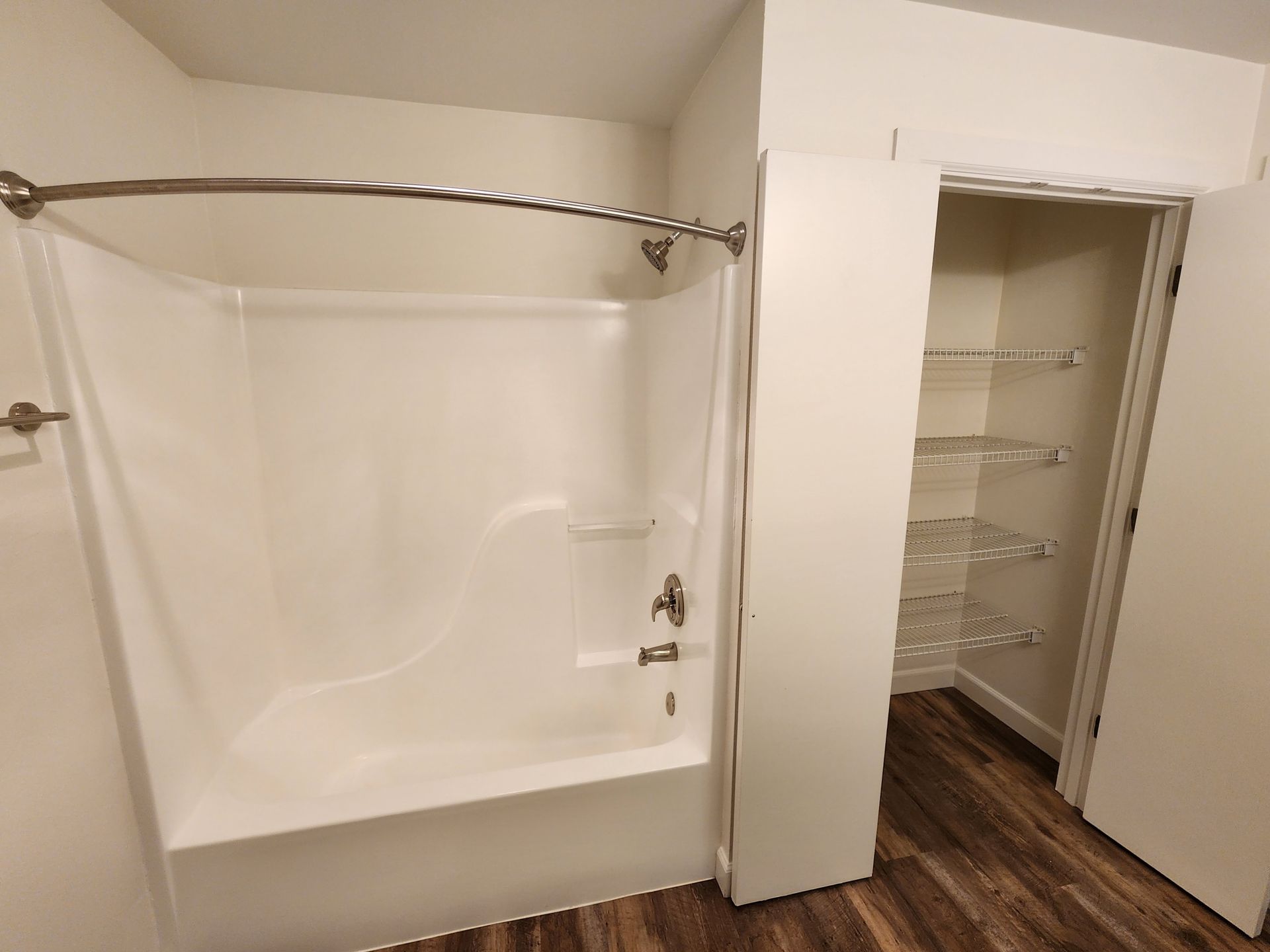 Bathroom with white bathtub, shower rod, and built-in closet with shelves; wood-look floor.