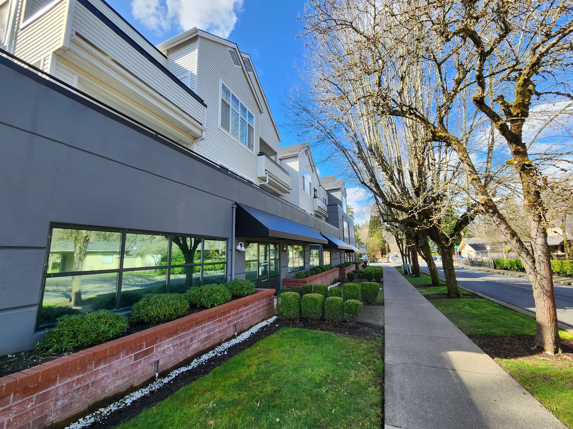 Building with storefront windows and a black awning, sidewalk, green grass, and trees under a blue sky.