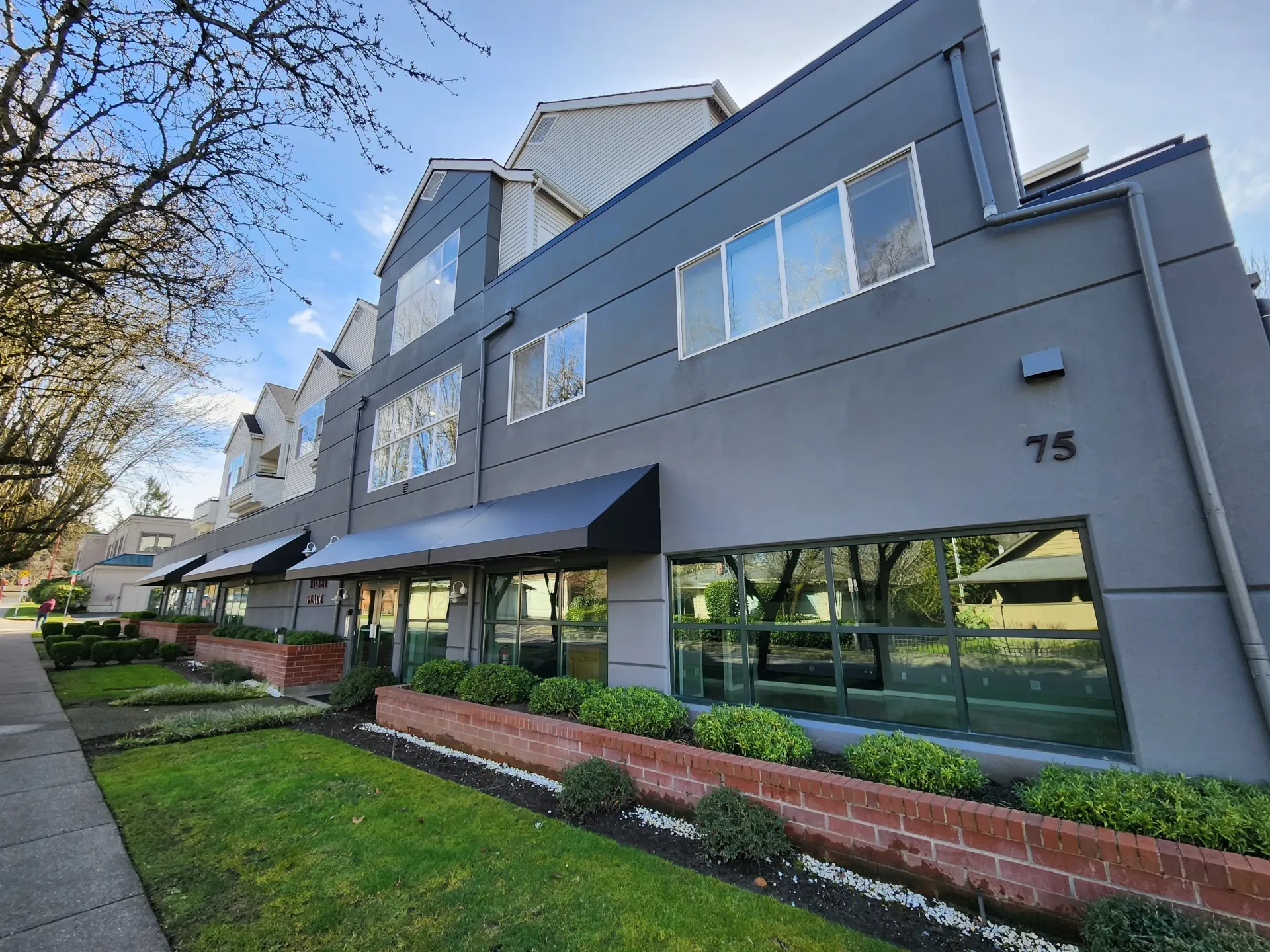 Gray commercial building with large windows, awnings, and landscaped beds; number