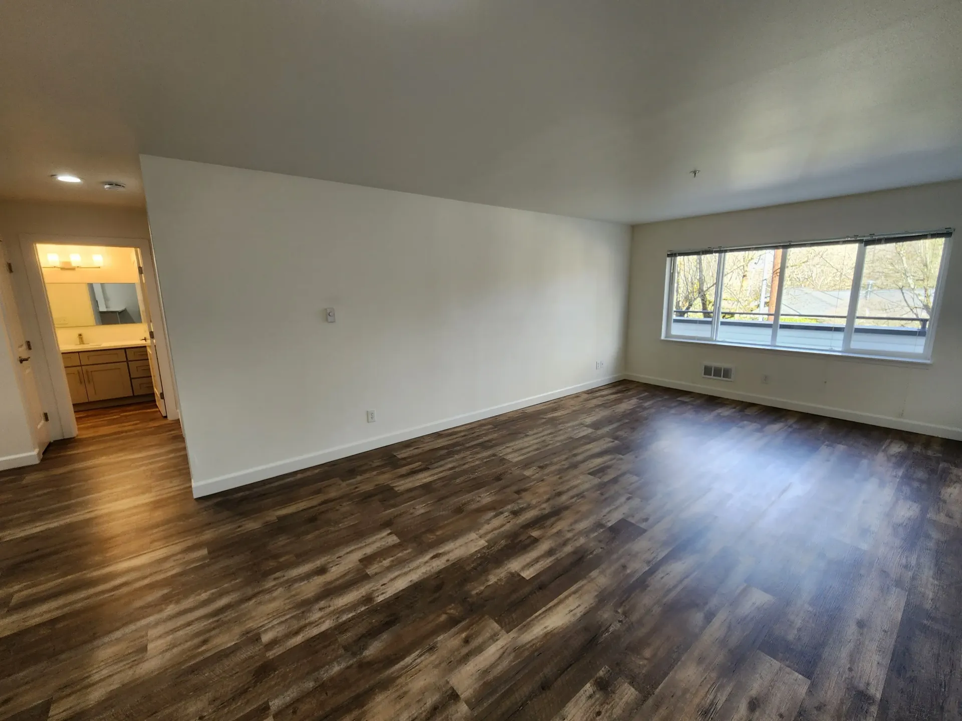 Empty room with wood-look flooring, white walls, and a window overlooking greenery. Bathroom visible through an opening.