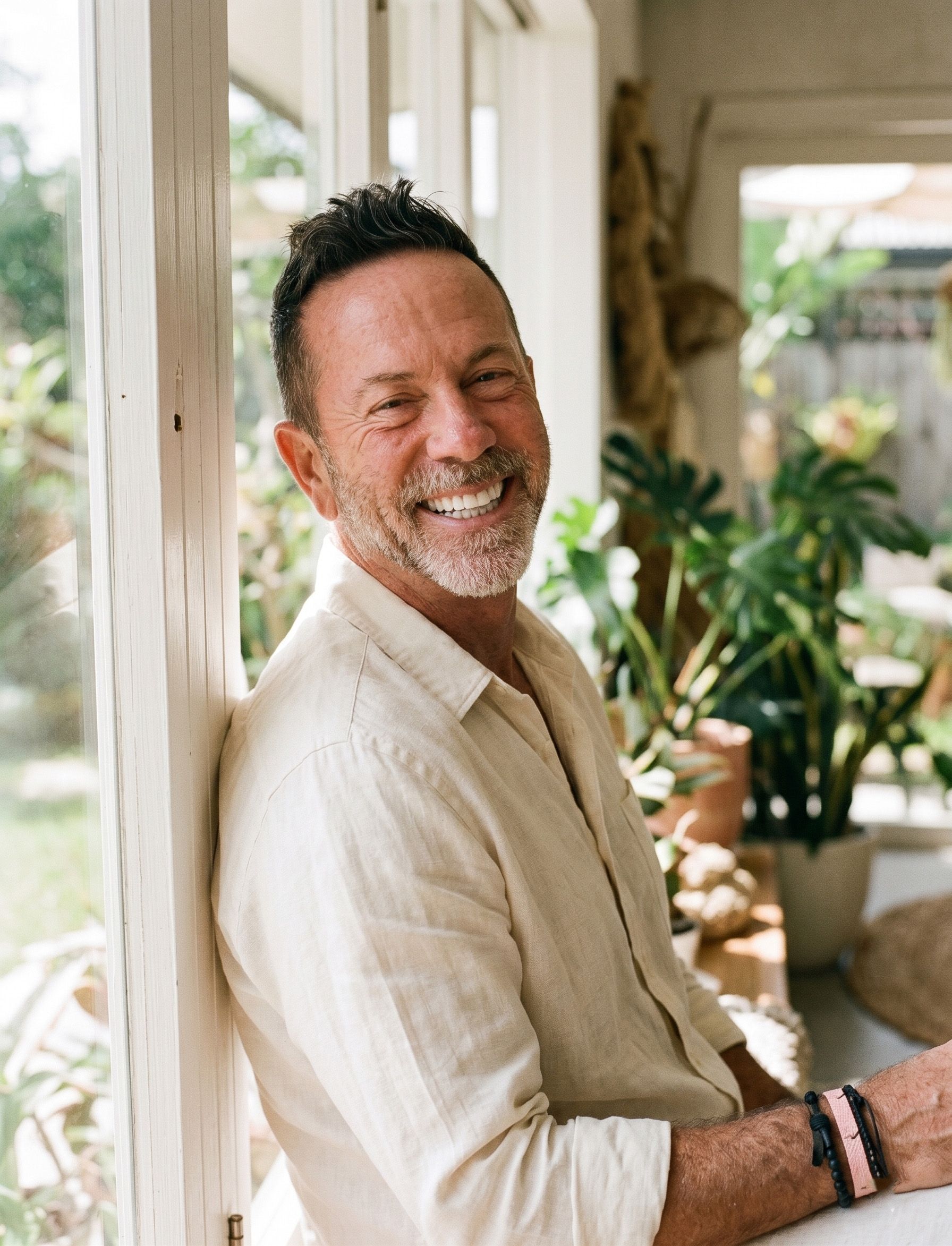 Smiling man in a beige shirt standing in a bright indoor space with potted plants.