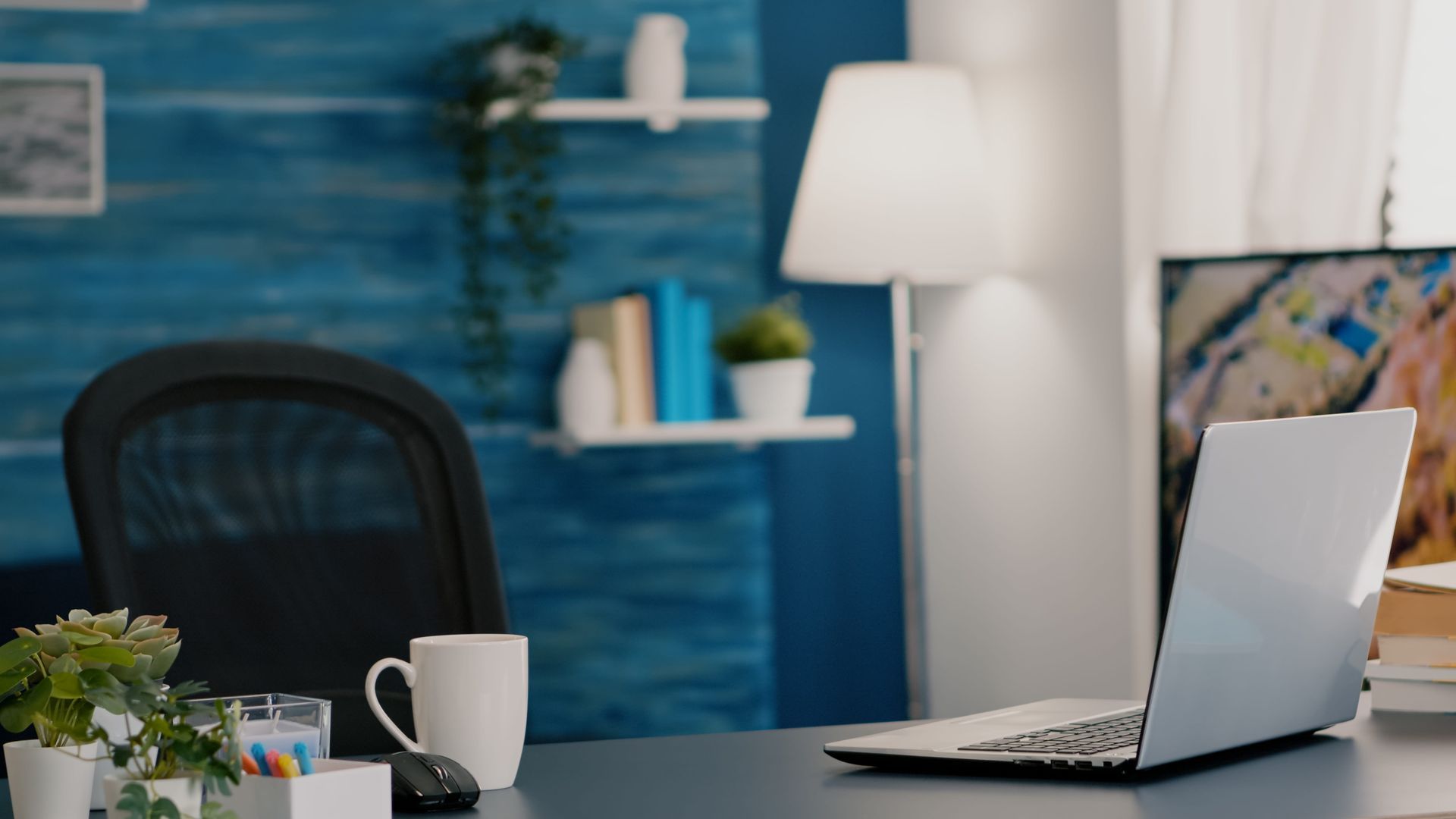 Modern office desk with laptop, coffee mug, and chair in a blue-walled workspace