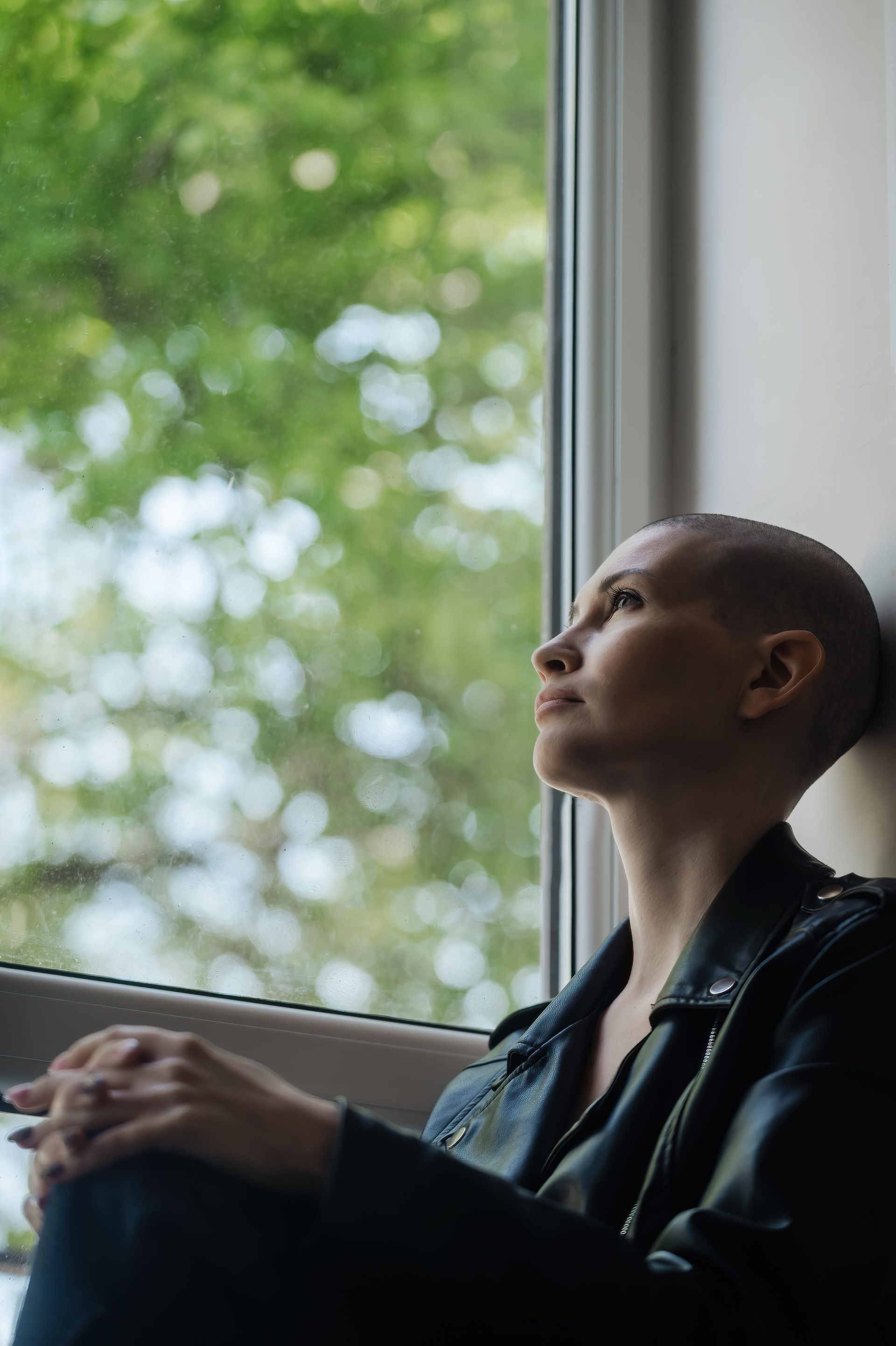 Person in a black jacket sitting by a window, gazing outside at the trees.