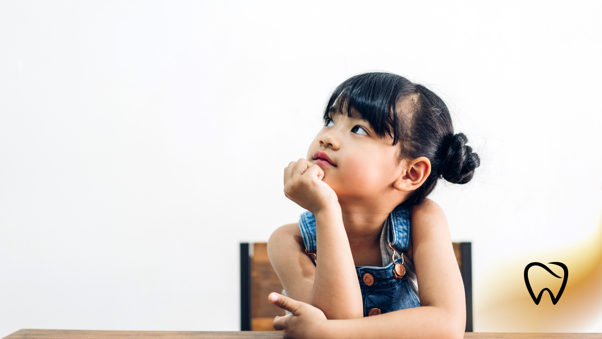 Young child with a thoughtful expression, looking upward, resting chin on hand at a table.