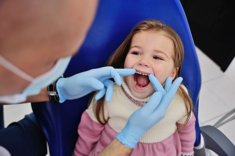 A dentist examines a young girl's teeth in a dental chair. The girl smiles, showing her mouth.