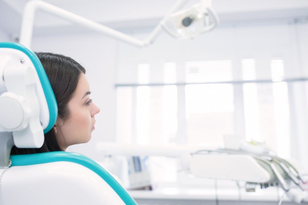 Woman in dentist chair, looking right. Teal and white chair in a bright dental office.