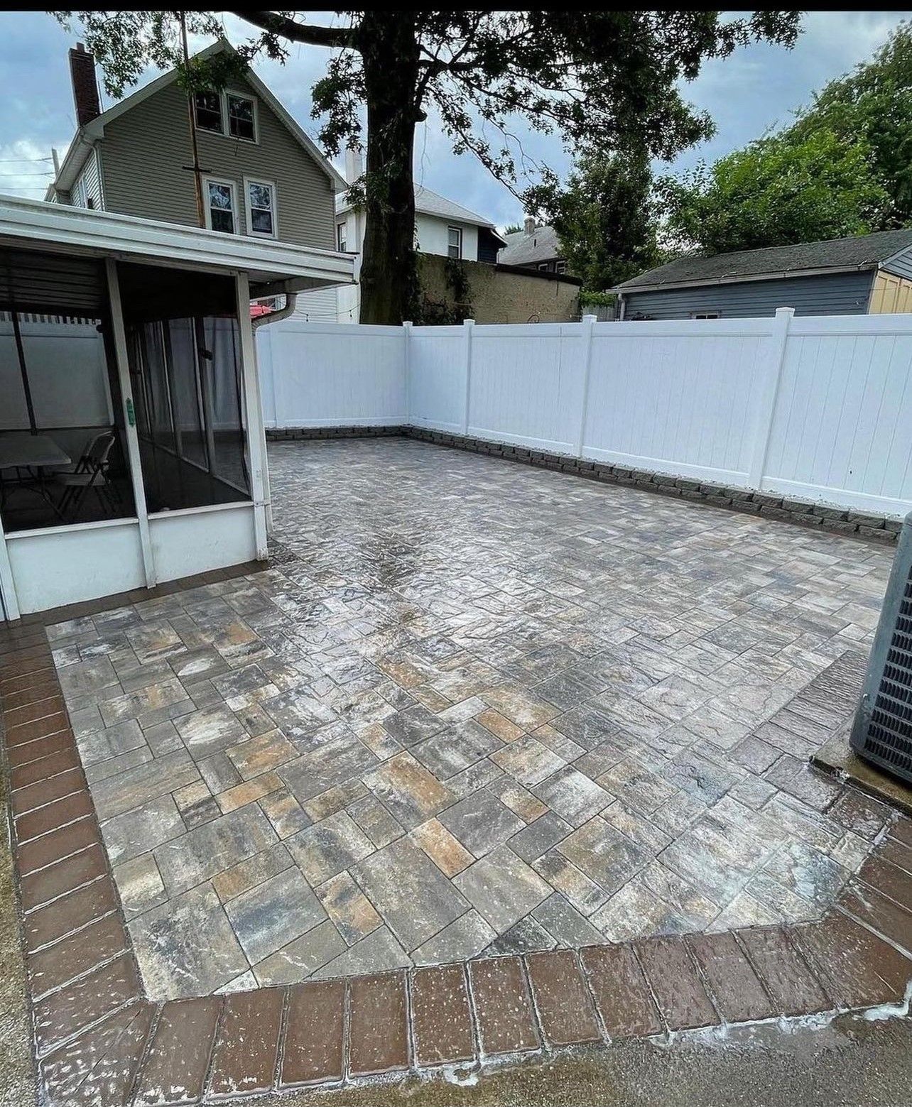 A backyard with a brick patio and a white fence.
