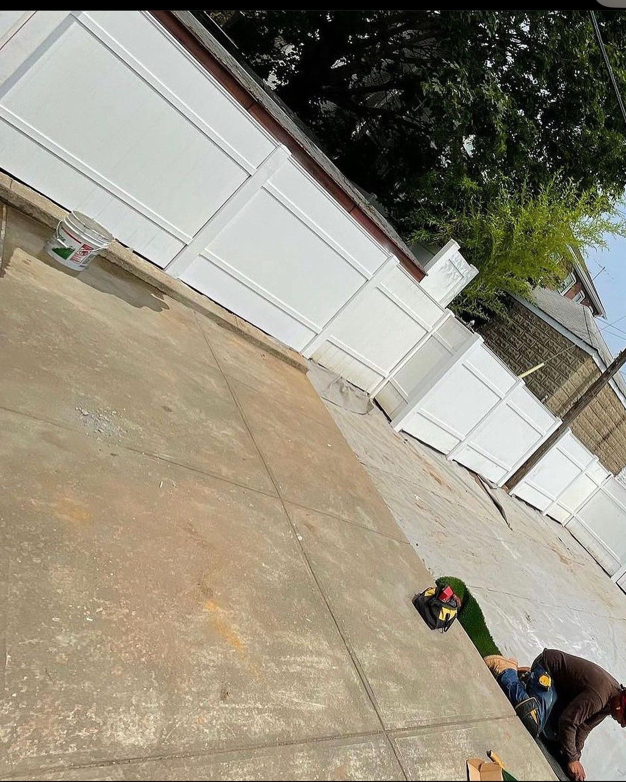 A person is standing on a sidewalk with a skateboard in front of a white fence.