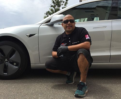 Man kneeling by a silver Tesla car wearing sunglasses, black gloves, and a black shirt.