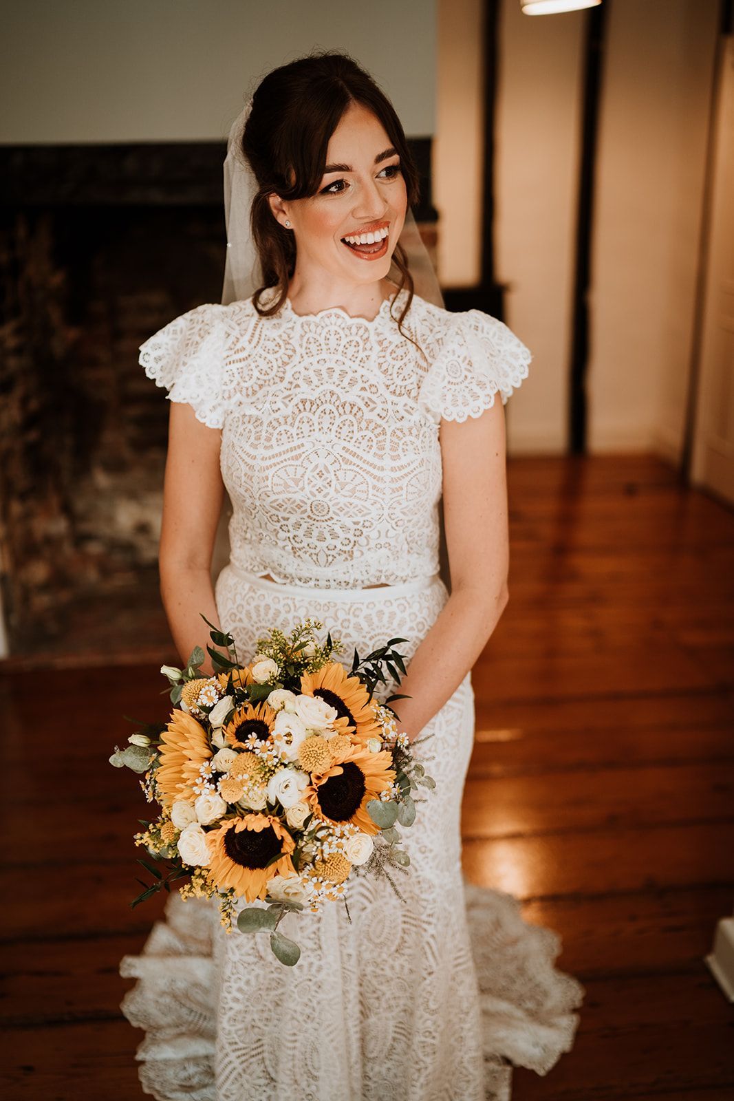 Smiling bride in a white lace dress holding a bouquet of sunflowers and white flowers indoors