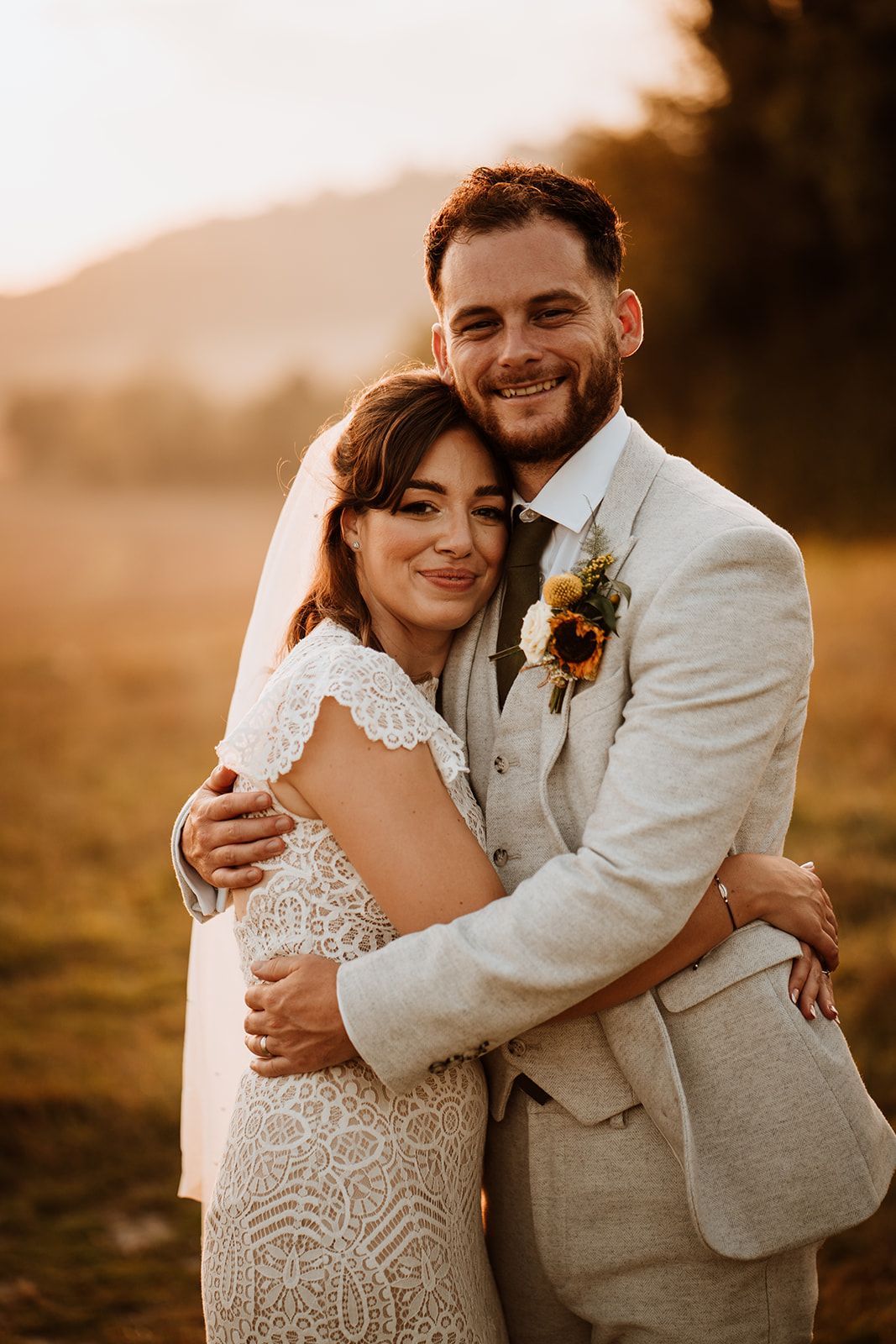 Smiling couple embracing outdoors at sunset, the woman in a white lace dress and the man in a light suit.
