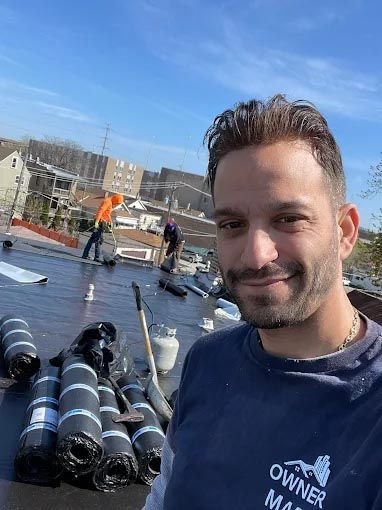A man is standing on top of a roof with rolls of roofing material.