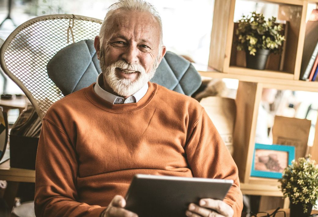 An elderly man is sitting in a chair holding a tablet computer.