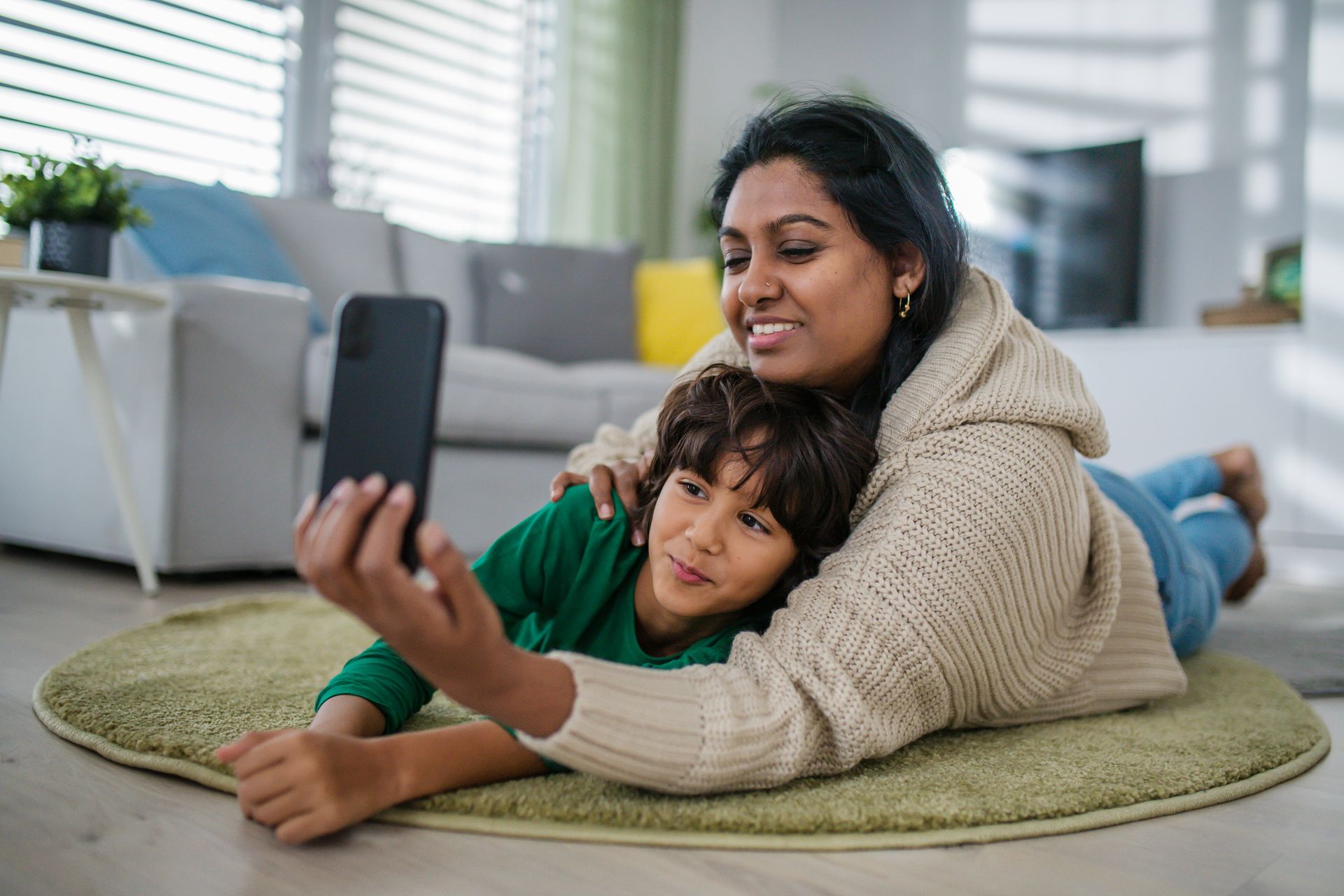 A woman and a child are laying on the floor taking a selfie with a cell phone.
