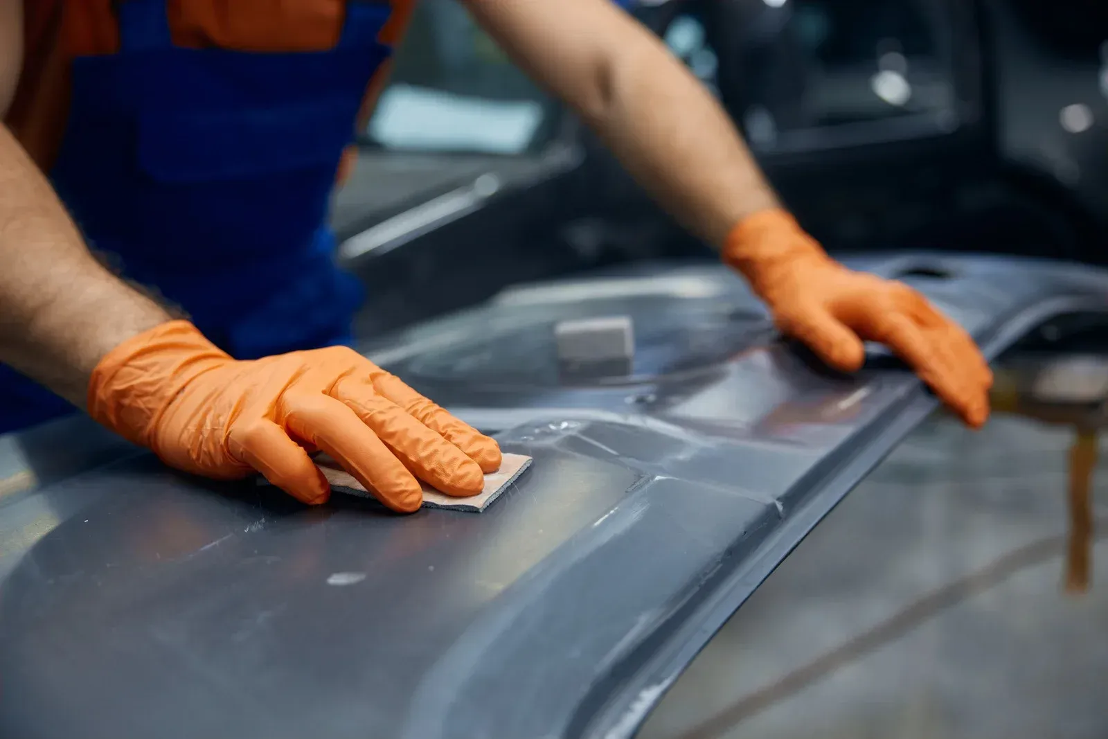 Person in orange gloves sanding a car part, wearing blue overalls.