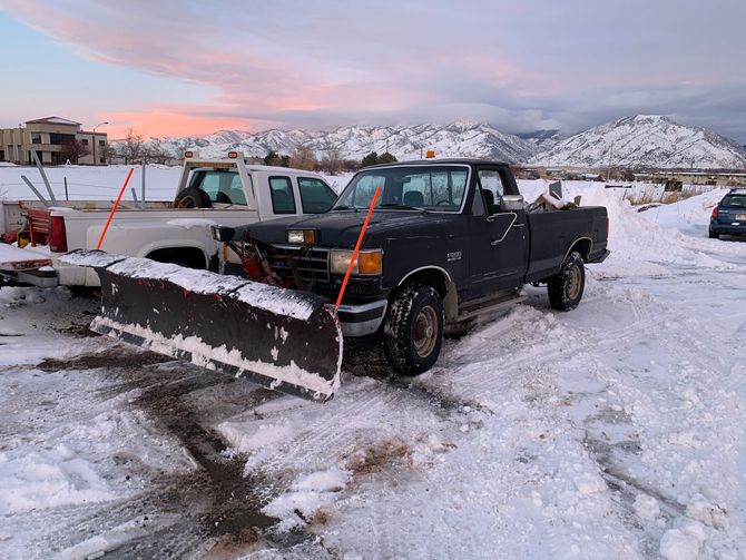 Black pickup truck with snowplow parked in a snowy lot with mountains in the background