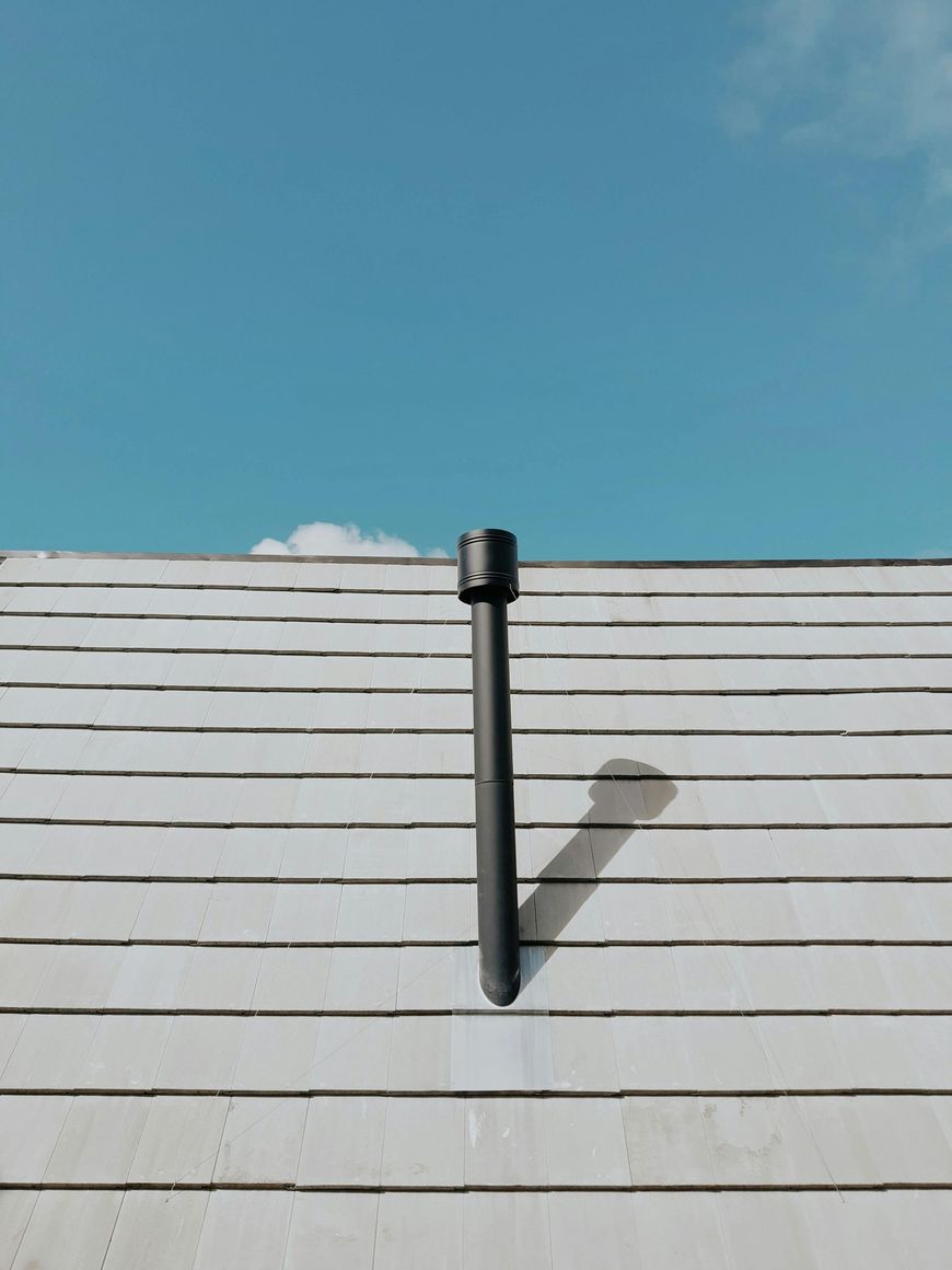 A black vent pipe protrudes from a light-colored, horizontal-patterned roof against a clear blue sky.