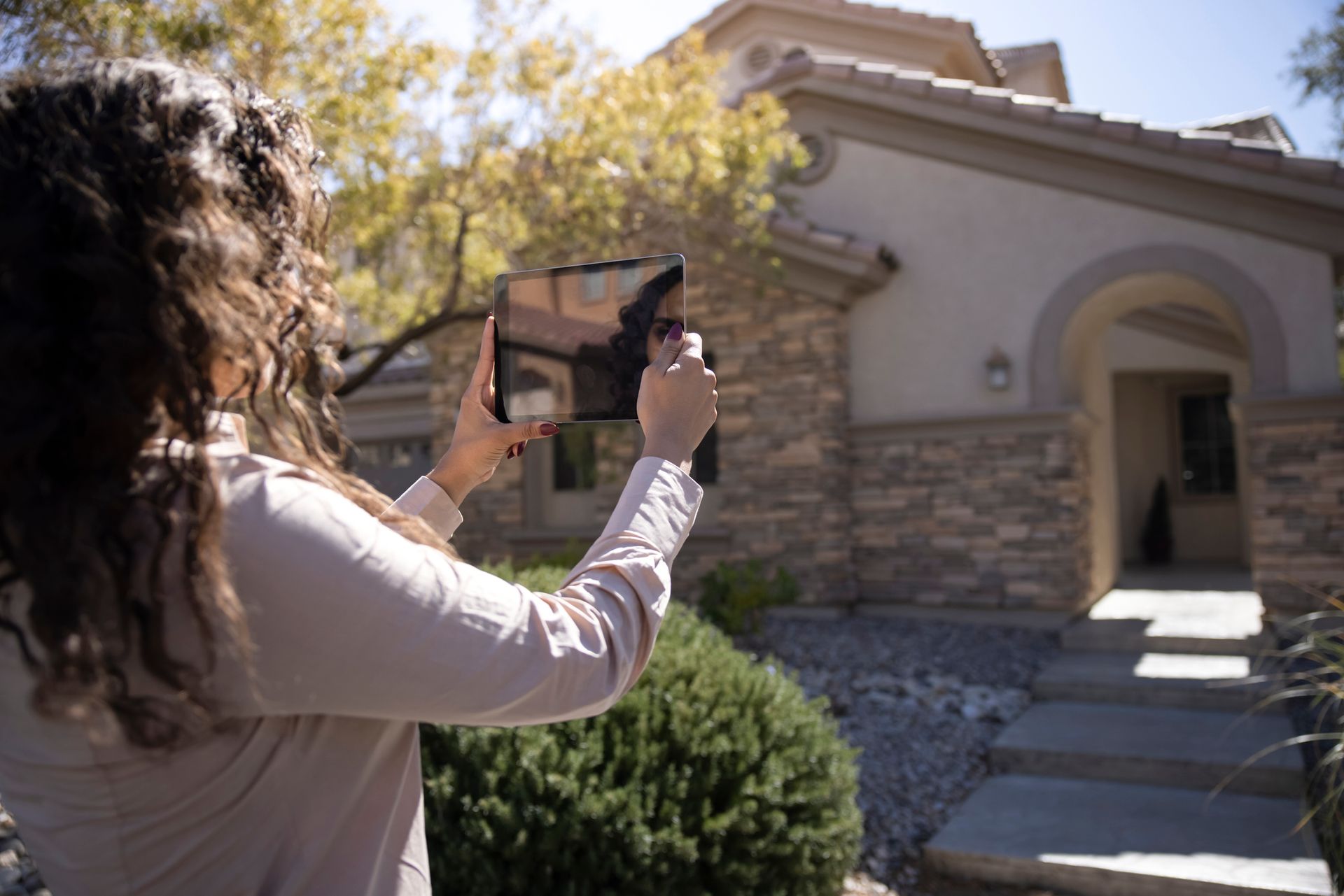 A feminine real estate broker is outside a home taking pictures with a tablet on a sunny day.