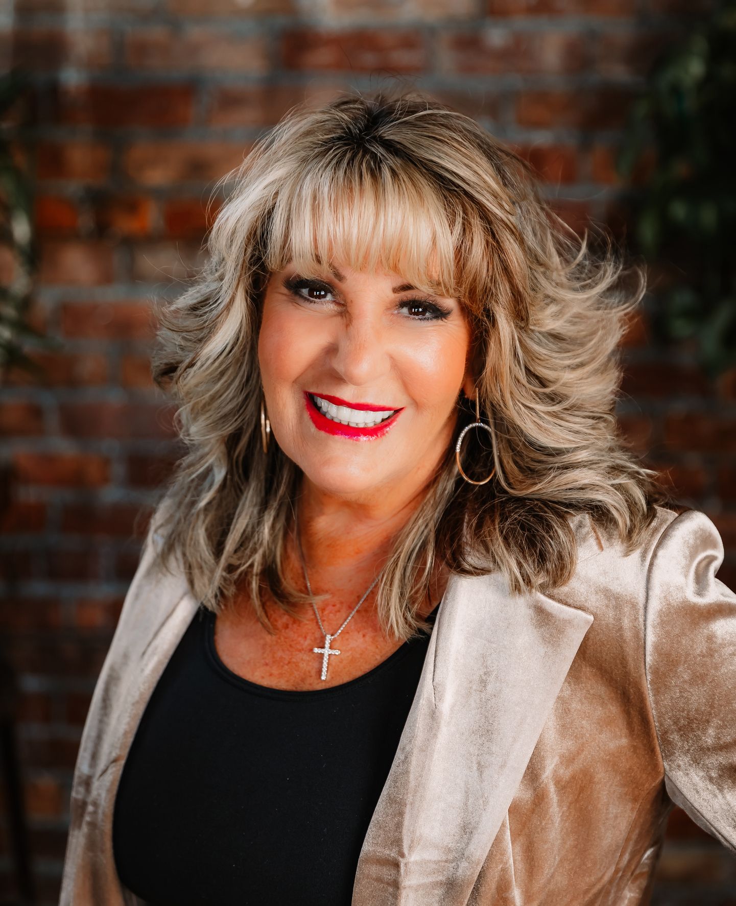 Jan Lazzara wearing a gold jacket and a black shirt is smiling in front of a brick wall.
