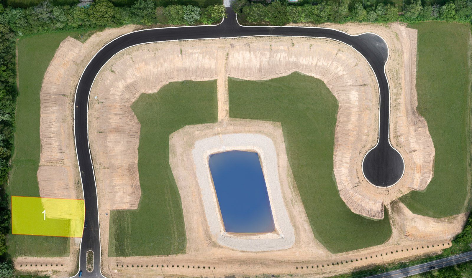 Aerial view of a new residential construction site with road and pond. Green grass and exposed earth are visible.