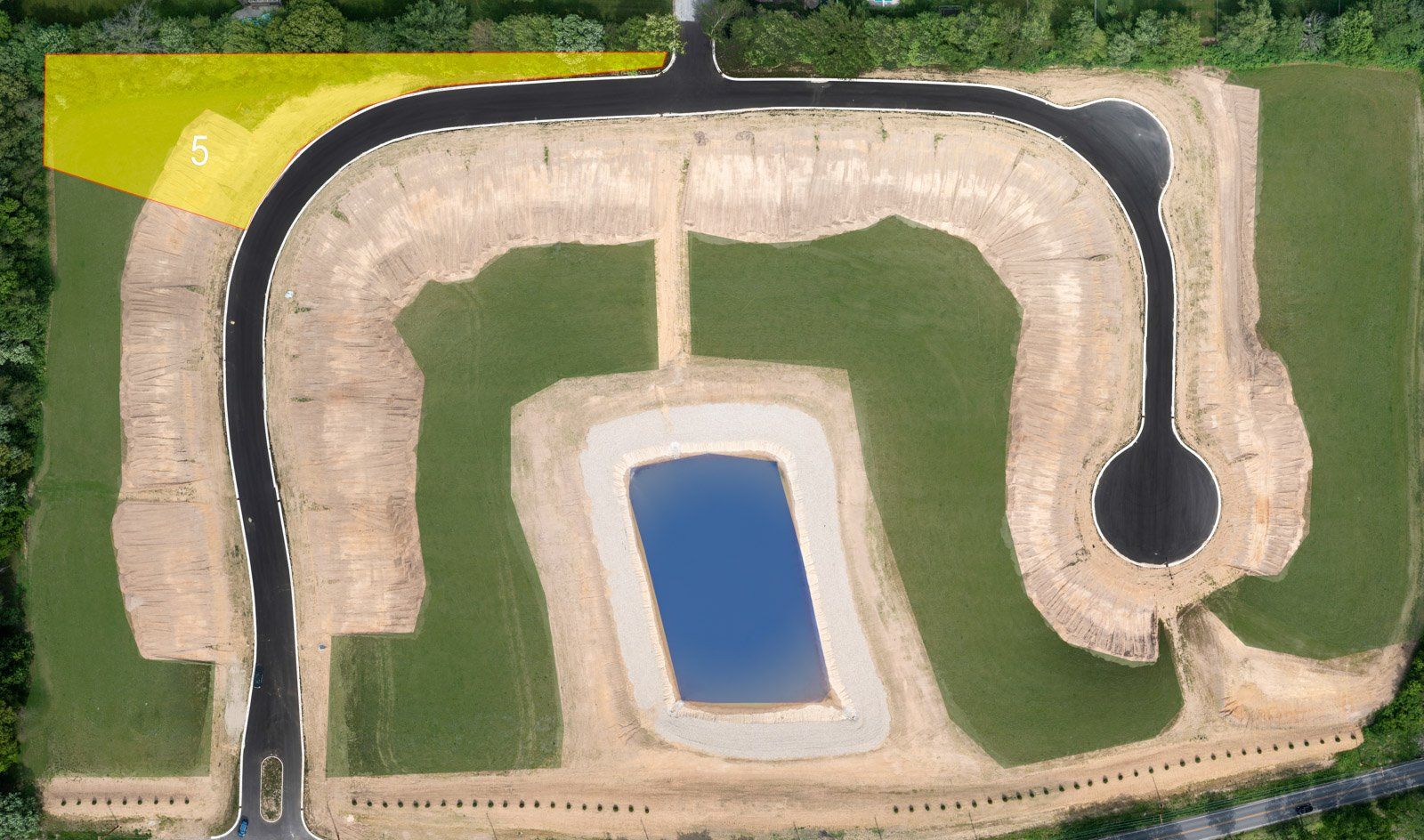 Aerial view of a new residential development under construction. Black roads curve around graded land with a central pond.