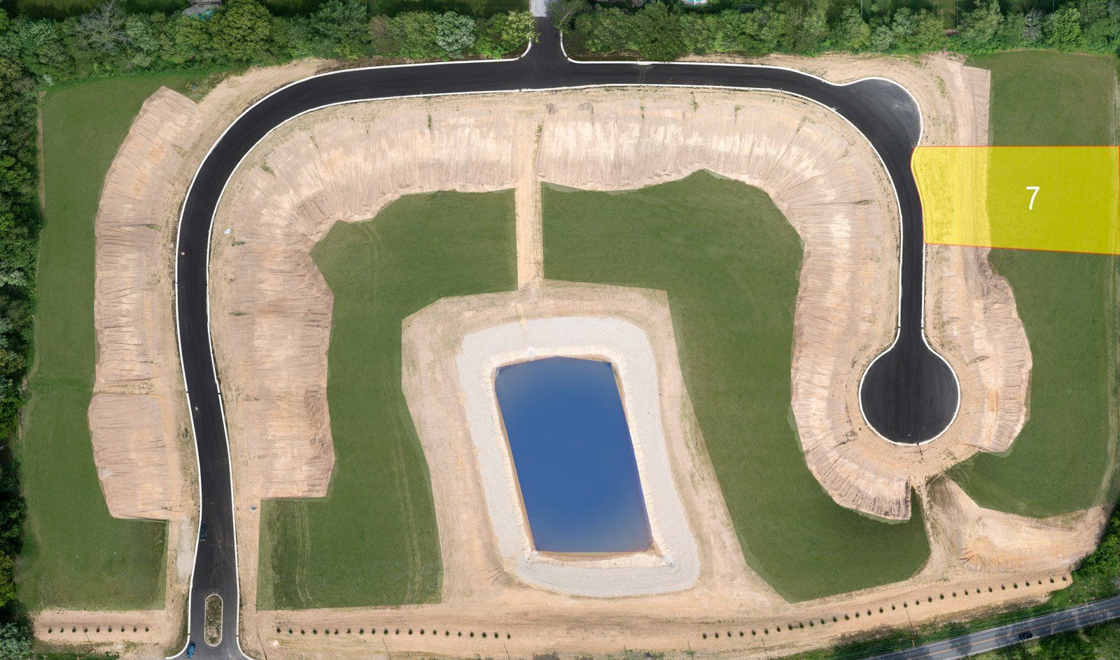 Aerial view of a housing development; asphalt roads, construction grading, a pond, and a marked lot (7) in yellow.