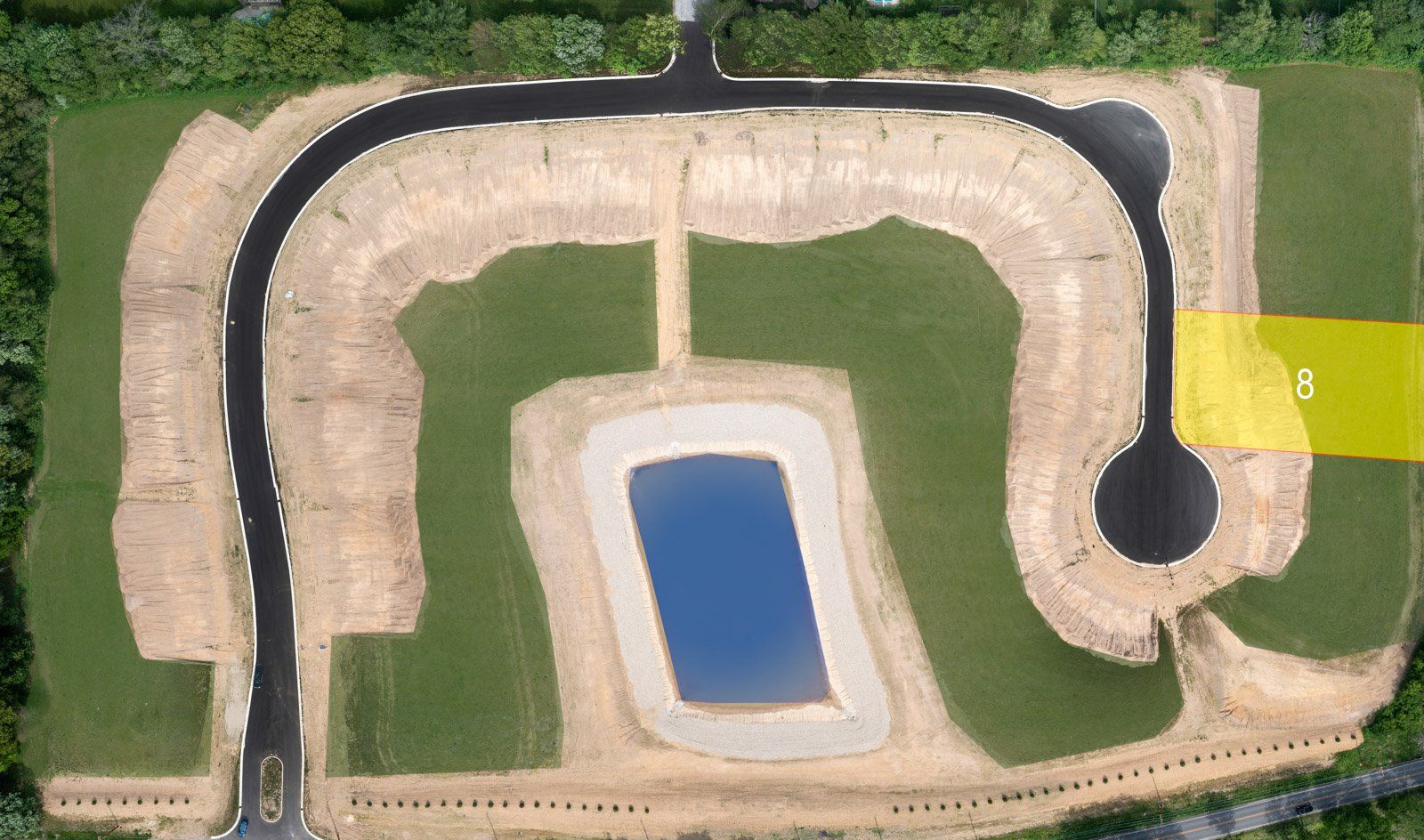 Aerial view of a new housing development with roads, a pond, and graded lots. Lot 6 is highlighted in yellow.