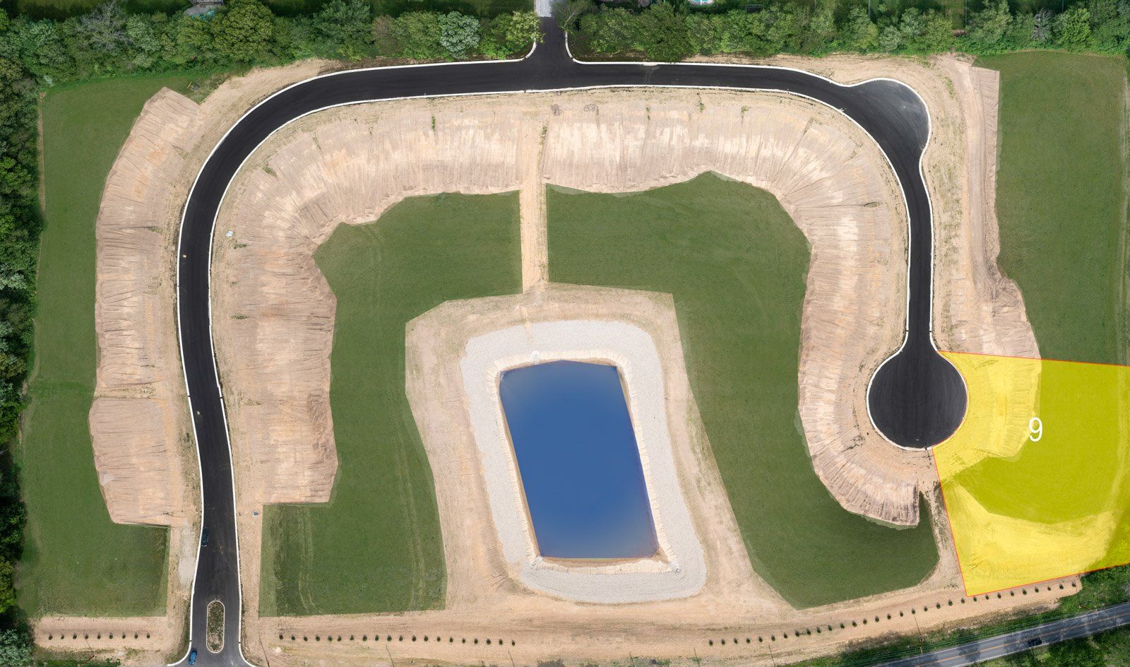Aerial view of a new housing development site with roads, a pond, and graded land. Green and brown colors dominate.