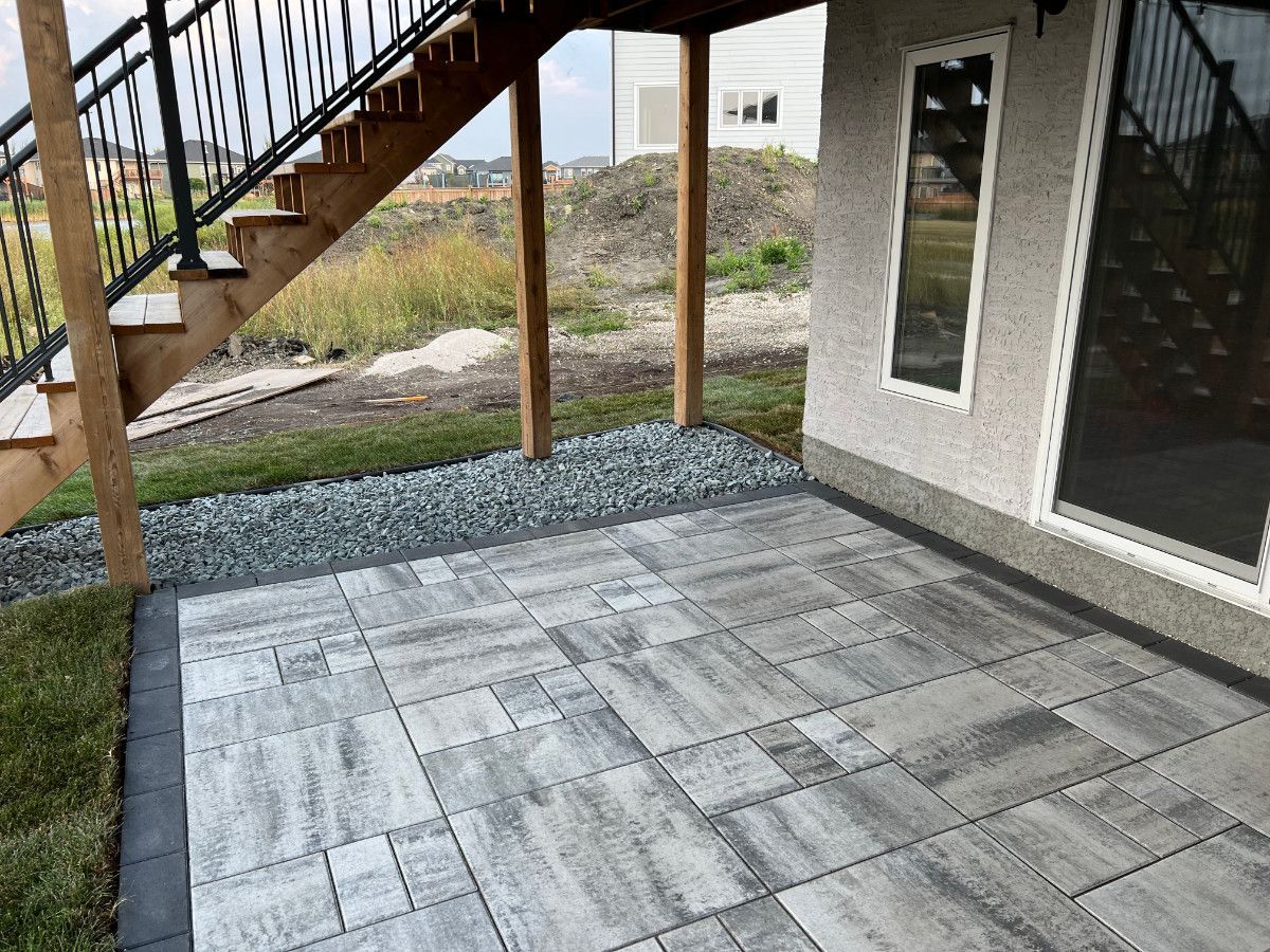 A patio made of light gray rectangular pavers with a dark border, situated under a wooden staircase against a house wall.