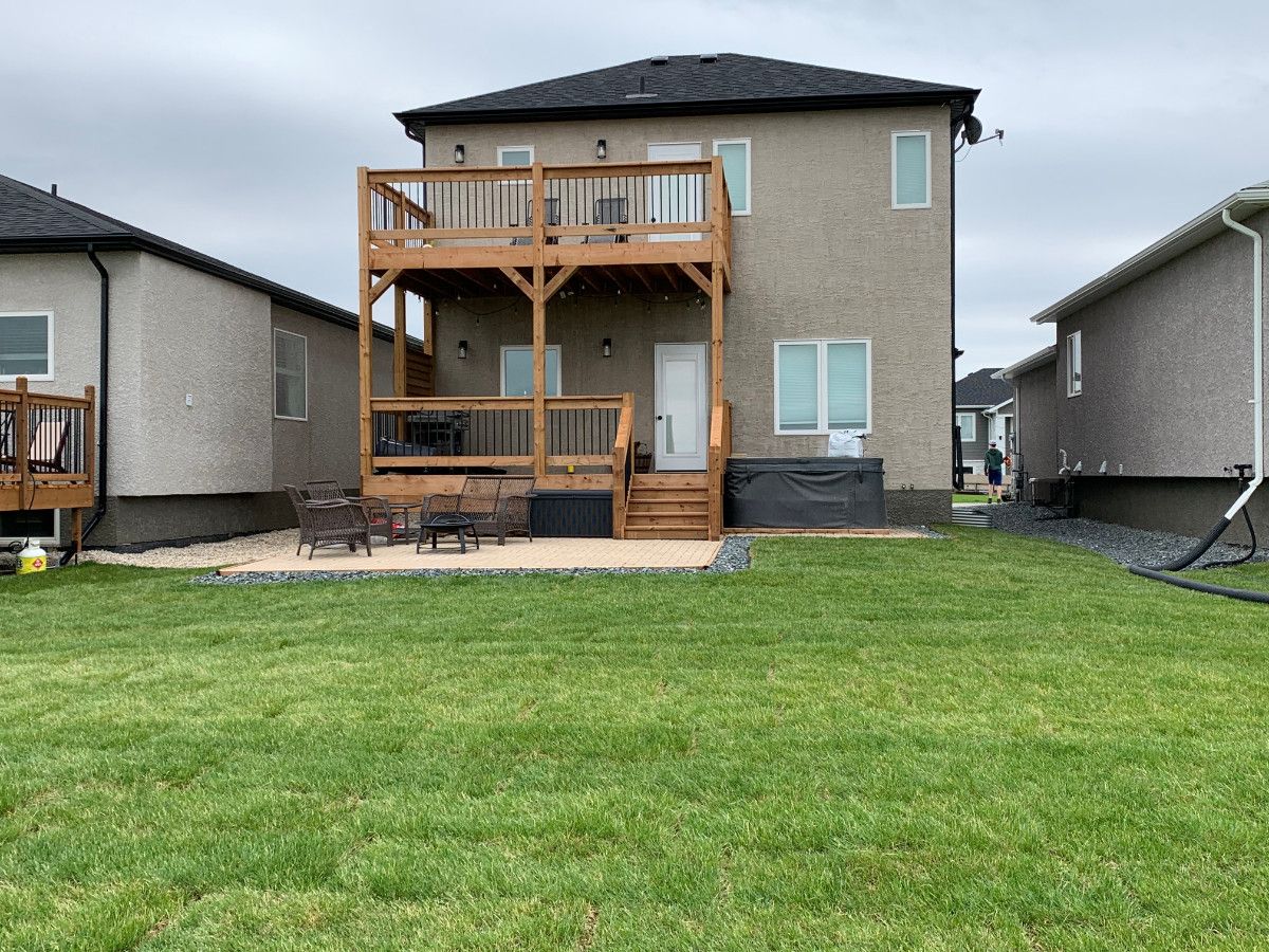 Back view of a tan two-story house with a wooden deck, a ground-level patio, and a grass backyard.