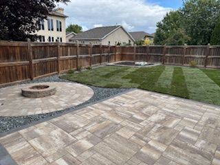 A backyard with a large paver patio, a circular stone fire pit, and a section of newly installed lawn inside a wood fence.