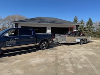 A blue and tan pickup truck parked in front of a garage, towing a utility trailer loaded with landscaping equipment.