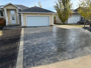 A residential driveway with gray and black patterned brick pavers, bordered by a beige stone edge in front of a house.
