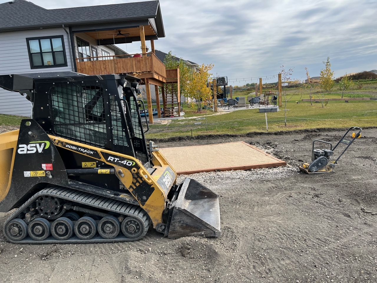 A yellow ASV skid steer loader sits on a construction site next to a leveled gravel patio base and a plate compactor.