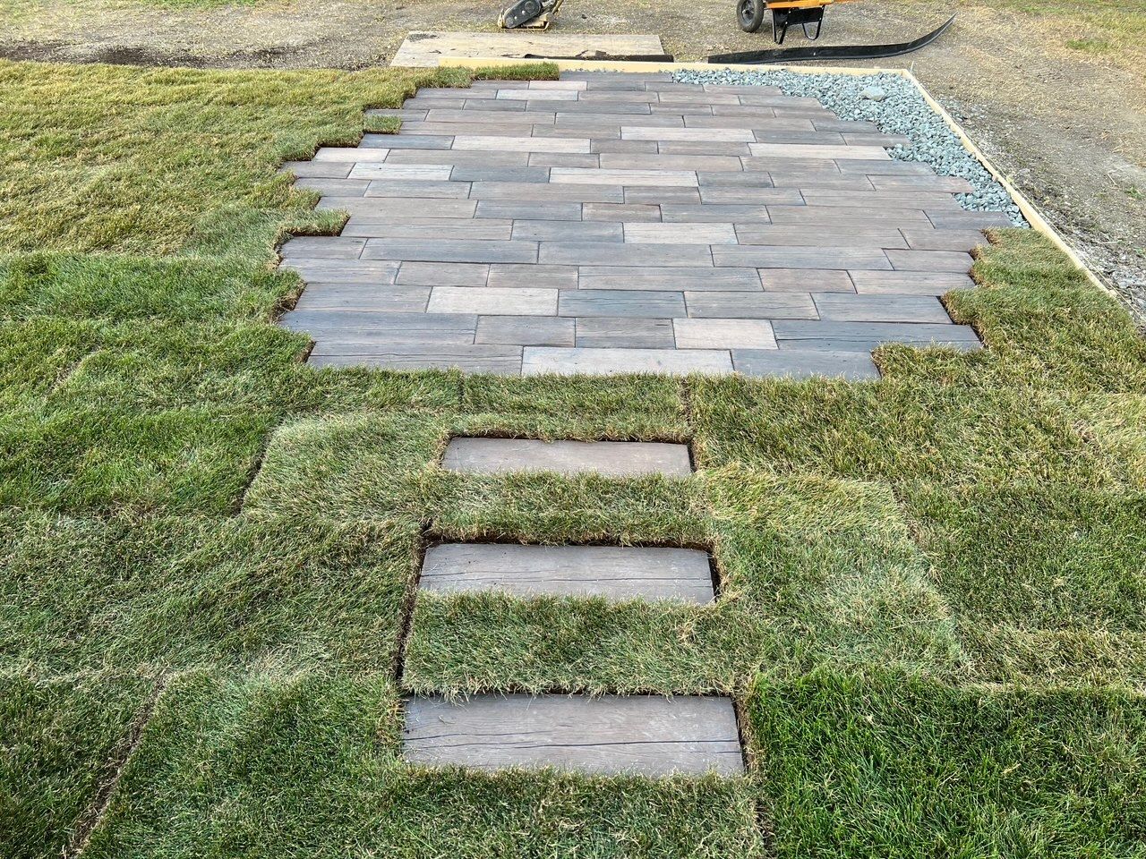 A patio of grey stone pavers connects to a walkway of three rectangular stones surrounded by freshly laid green sod.