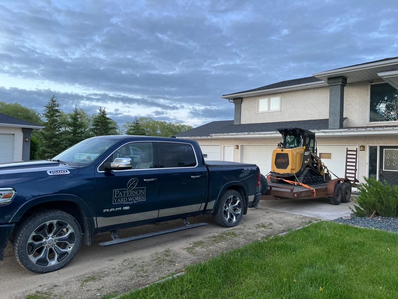 A dark blue pickup truck parked in a residential driveway, towing a trailer with a yellow skid-steer loader.