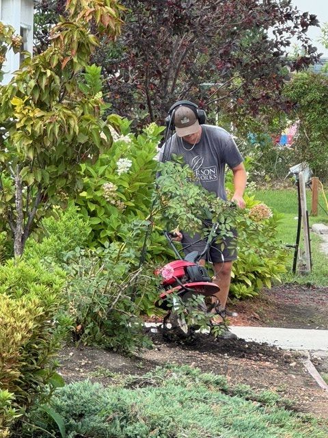 A person wearing earmuffs and a grey t-shirt uses a red tiller in a garden bed near a sidewalk.