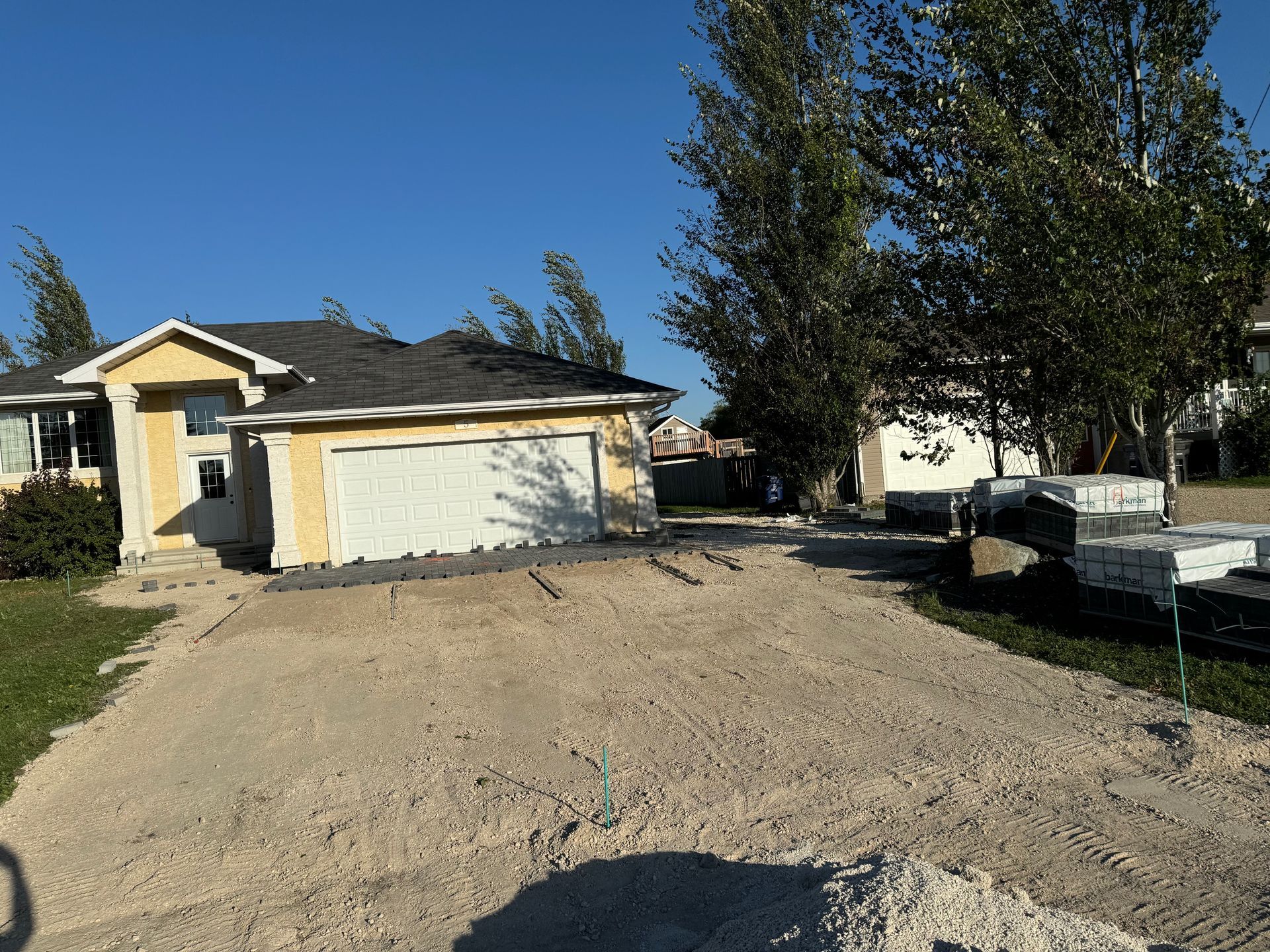 A yellow house with a white garage door, featuring an unpaved, gravel driveway under construction in a suburban setting.