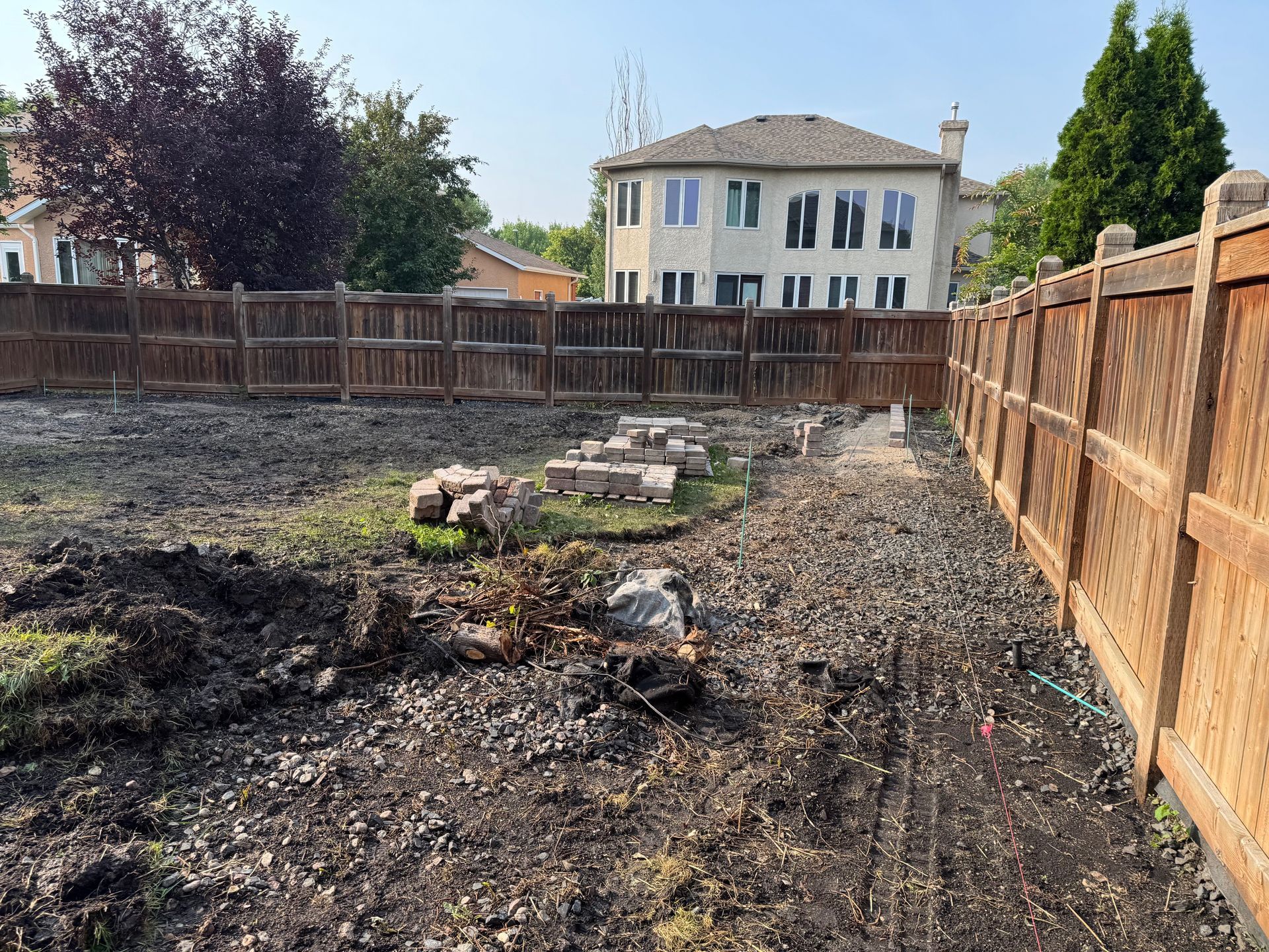 A backyard under construction with dark, churned soil, a wooden perimeter fence, and a large house in the background.