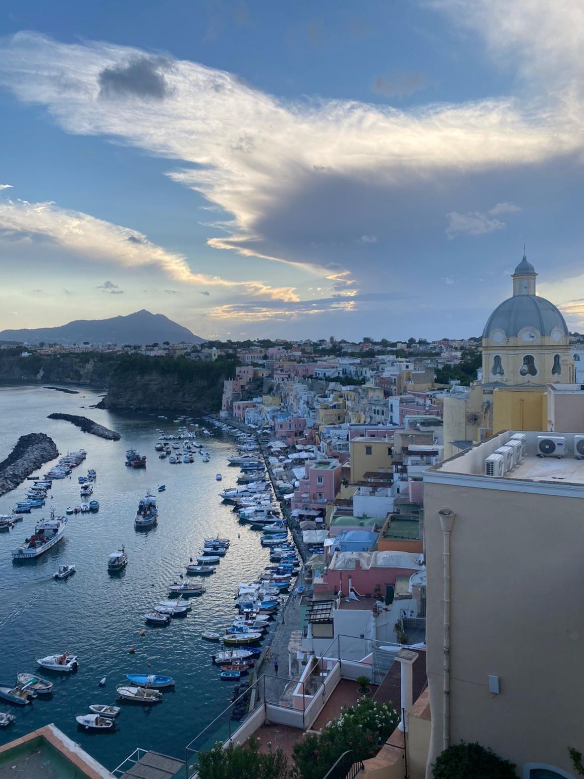 Città costiera che domina il porto, con edifici colorati, barche e una chiesa con cupola sotto un cielo parzialmente nuvoloso.