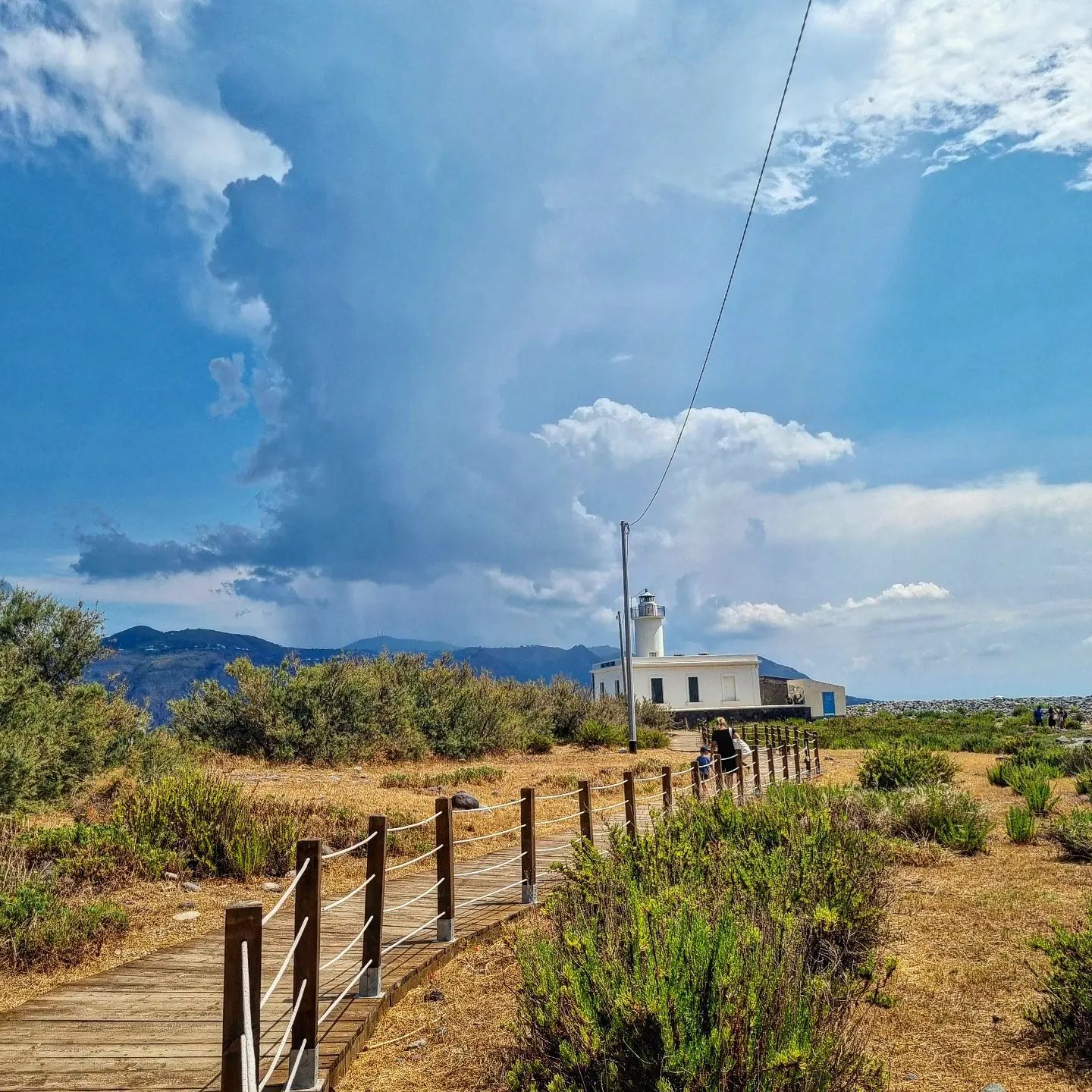 Passerella in legno che conduce a un faro bianco sotto un cielo nuvoloso. Cespugli verdi e montagne sullo sfondo.