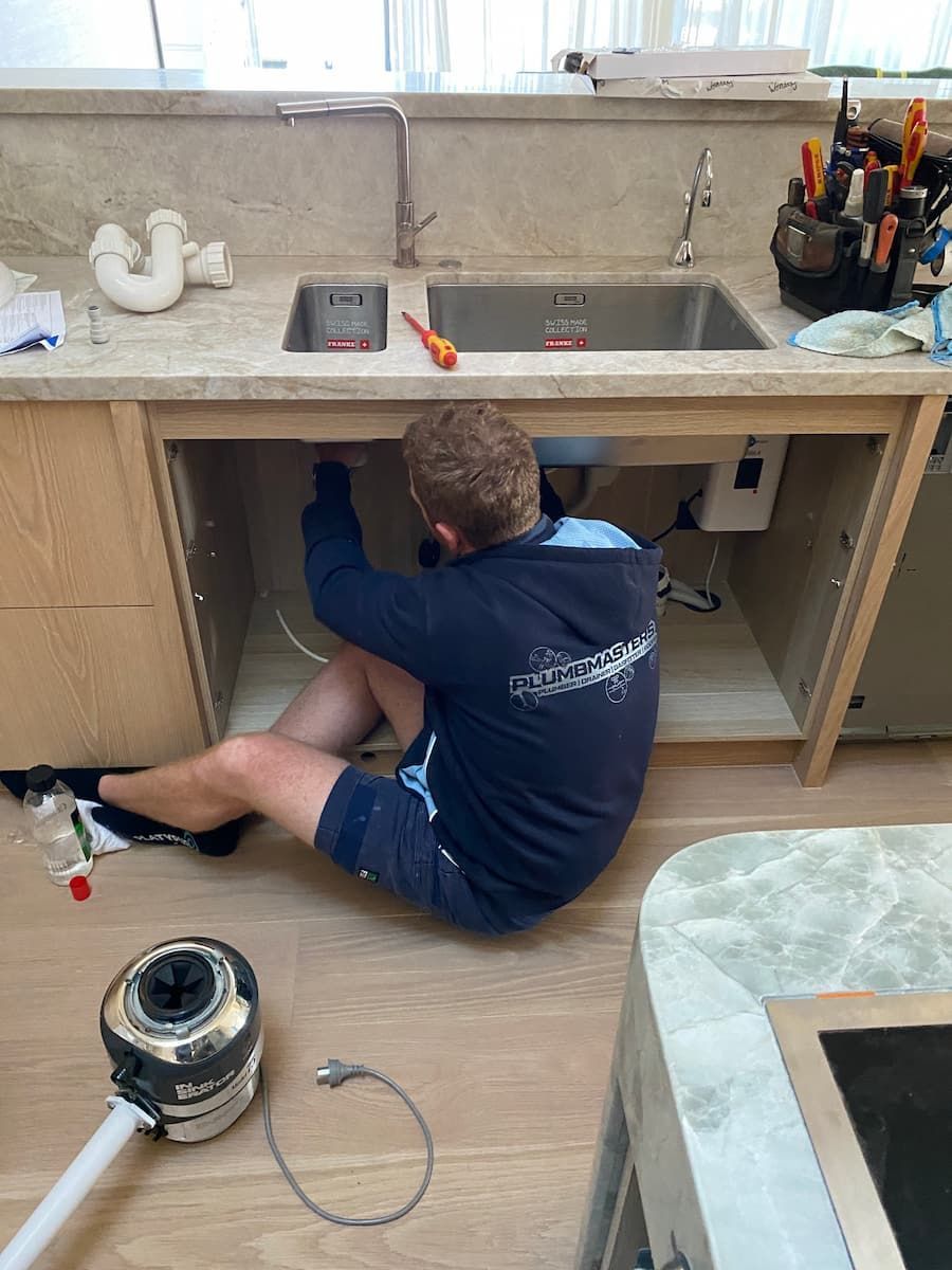 A Man Is Kneeling Under A Sink In A Kitchen — Plumbmasters In Burleigh Heads, QLD